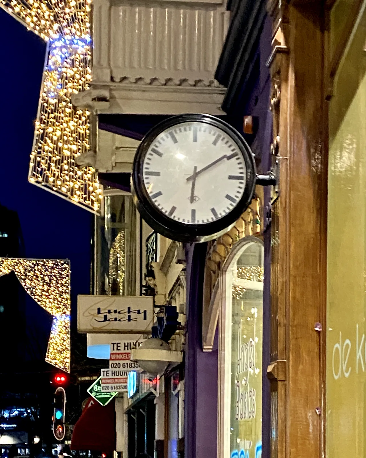 A clock attached to a facade in a colourful shopping street points at 9 minutes past 6.