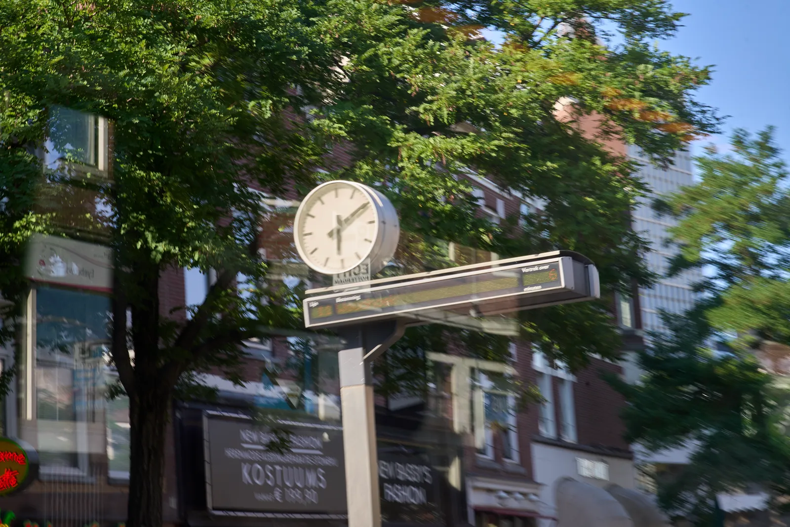 A clock on a pole, with a thin, wide digital display below it. The whole picture is a bit blurry, it seems like it’s taken from a moving vehicle. There are green trees and a brick building in the background. The clock points at 9 minutes past six.