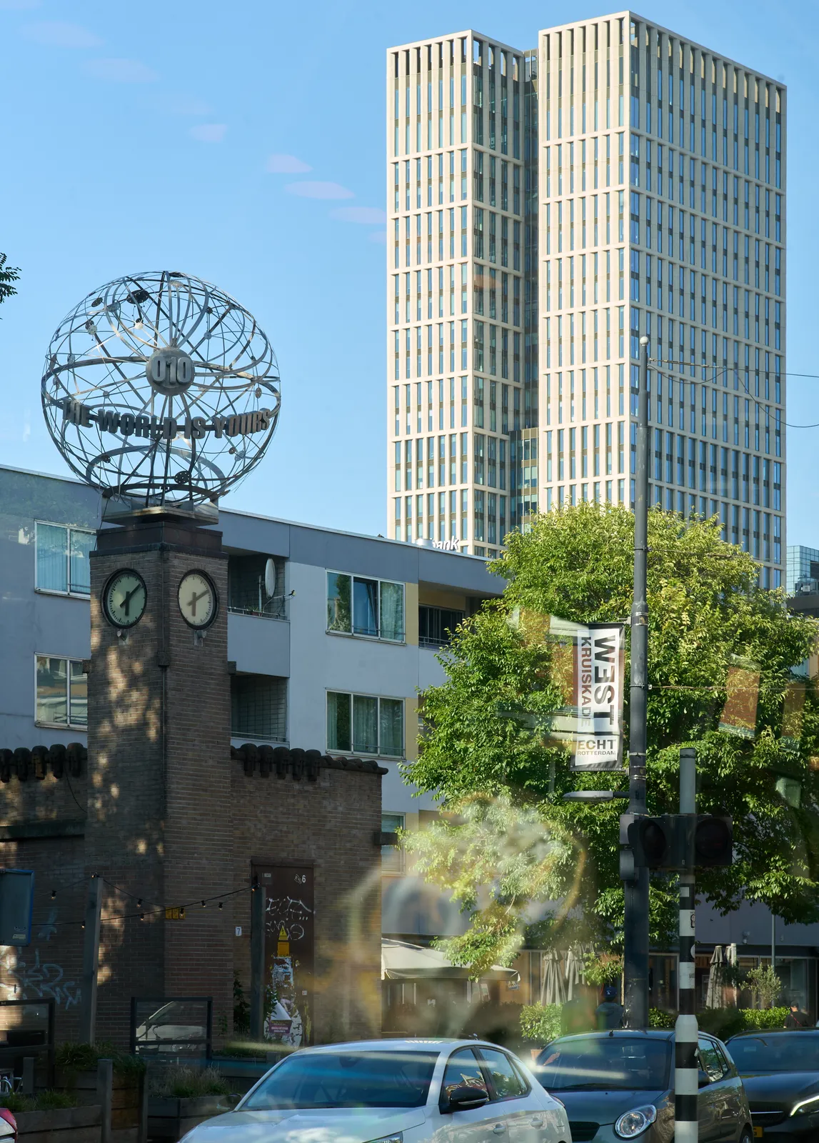 Two enormous skyscrapers in the background, against a bright blue sky. In the foreground there’s a brick building with a tower with clocks on it and a large globe on top with the words: The world is yours. This building stands on front of some impoverished apartments. There are two clocks visible on the tower. The one on the left points at eight minutes past six, the one on the right points at 10 past six.