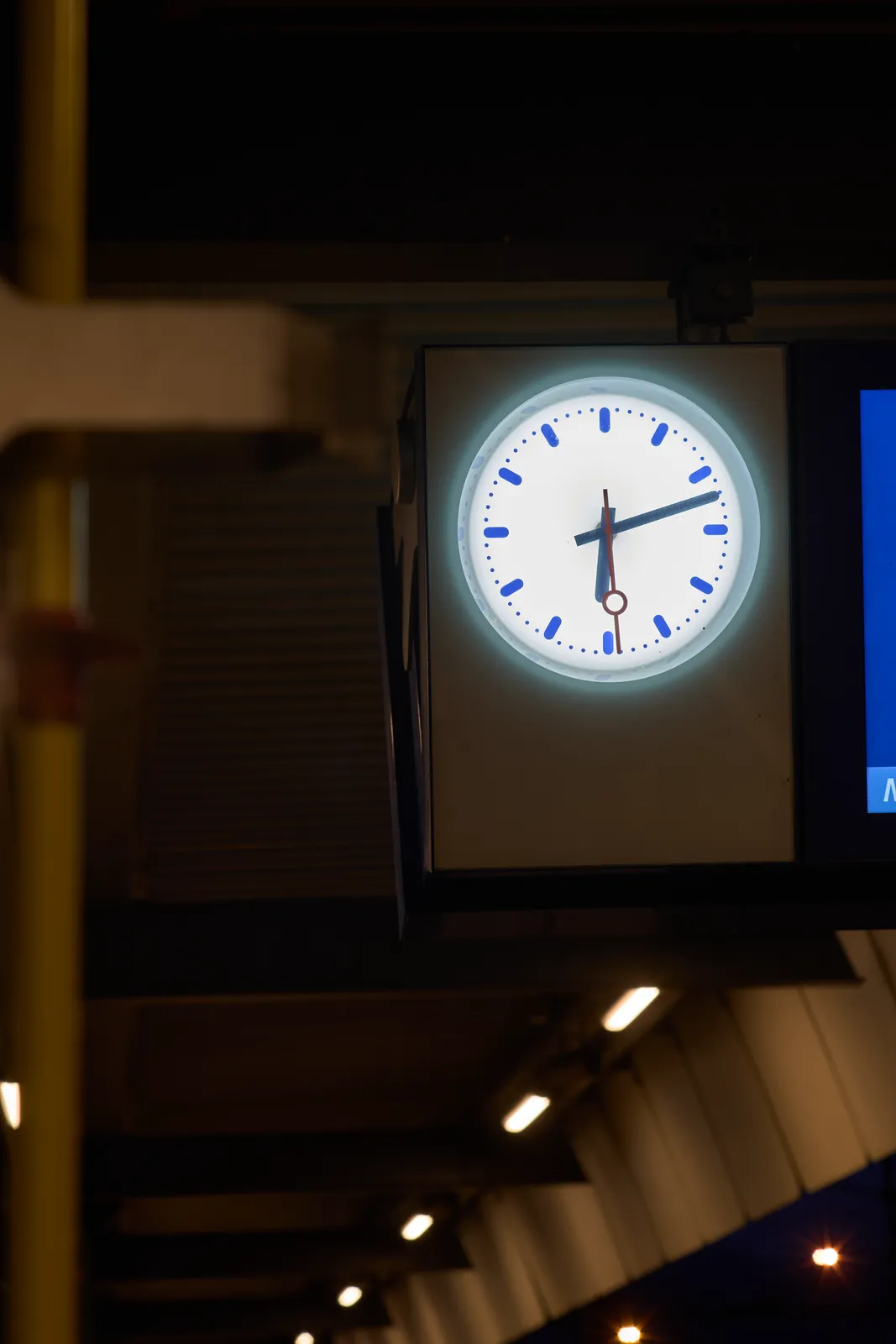 A lit station clock on a dark platform. It points at 12 minutes past six.