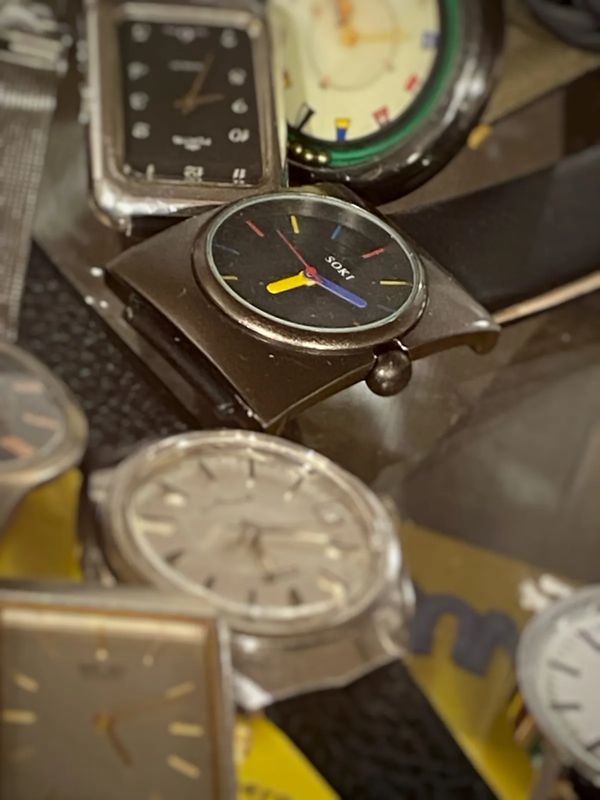 A few wrist watches on a table. One of them, a rather novel type of watch, is in focus and it points at 12 minutes past 6.