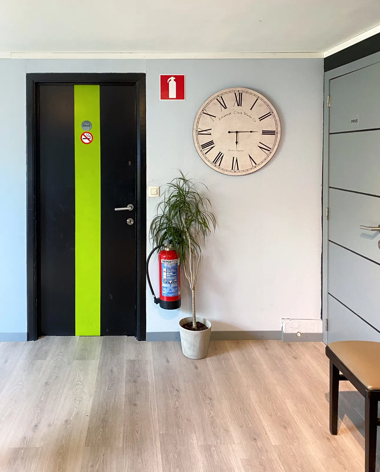 A black and green door with a non-smoking sign on it. Next to it there’s a plant that tries to cover a fire extinguisher, in vain. Higher up on the wall there’s a sticker that indicates that the fire extinguisher can be found here. Next to all this there’s a large clock that points at a quarter past 6.