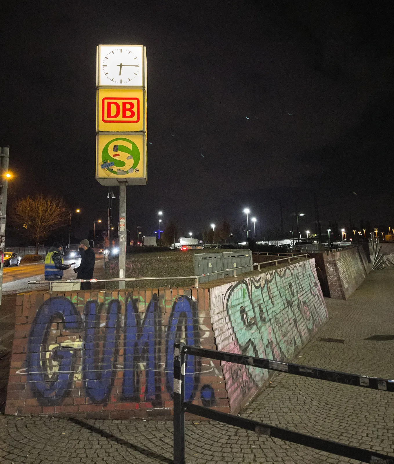 A lit clock on a pole, standing on some nicely curved brick walls. There are two large logos below the clock, one of these logos is from the german railways. The clock points at a quarter past 6.