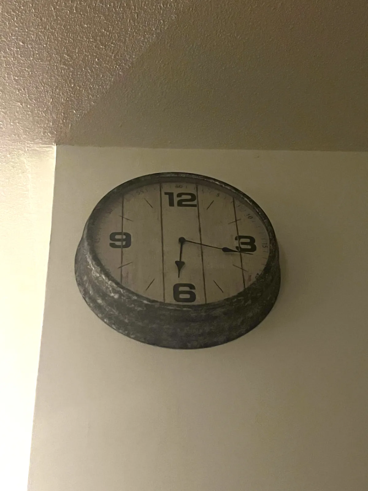 A clock is attached to a shadowy side of a wall. Light beams past the corner. The clock seems to be made from some sort of metal, and it looks old and used. On the face only the numbers 12, 3, 6 and nine are displayed. The hands point at 17 minutes past 6. 