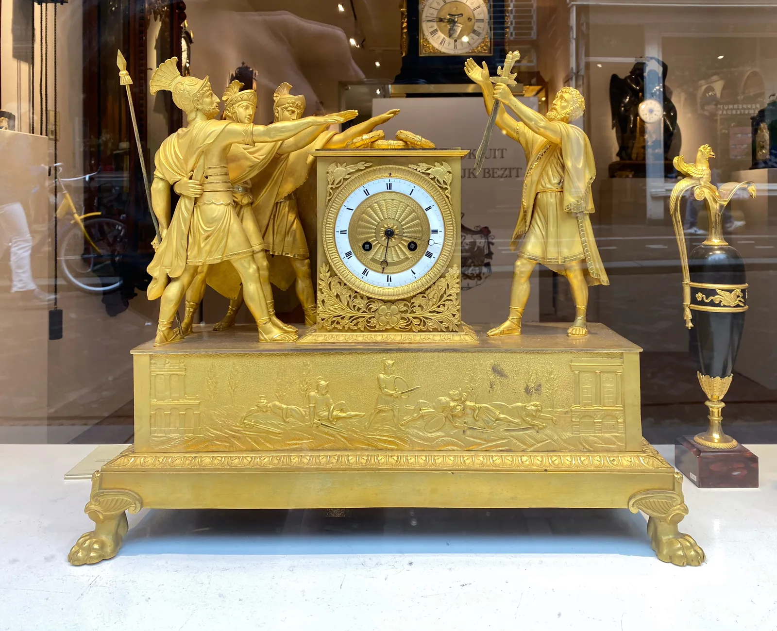 Some statues of ancient Roman soldiers pointing at a statue of a man holding a paper at the other side of this golden clock. It points at  17 minutes past 6.