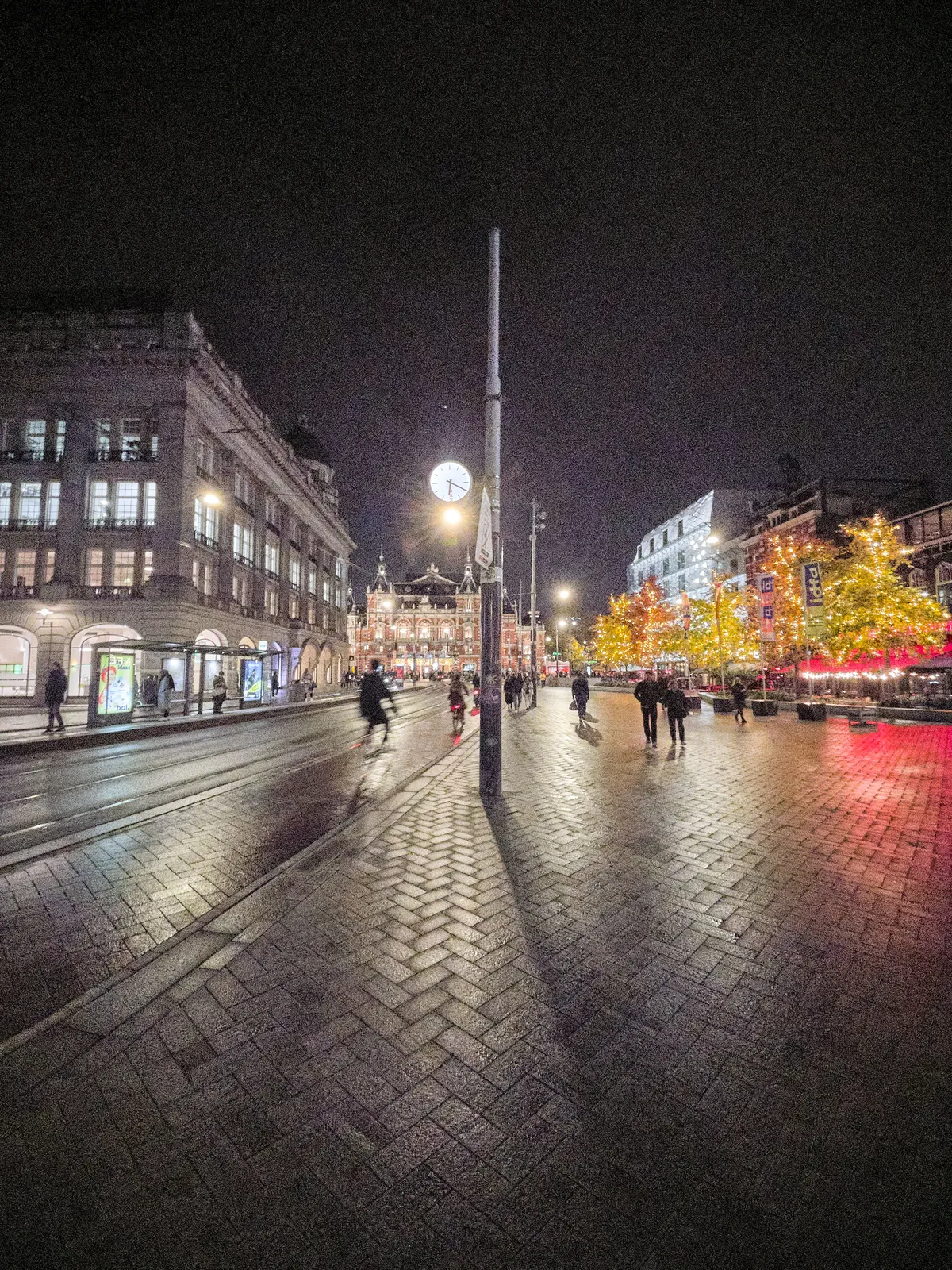 A clock attached to a pole, right in the center of an image of the Leidse Square in Amsterdam at night. Large buildings are lit, people are walking and biking, and everything is reflected in the wet street. The clock points at 20 past 6.