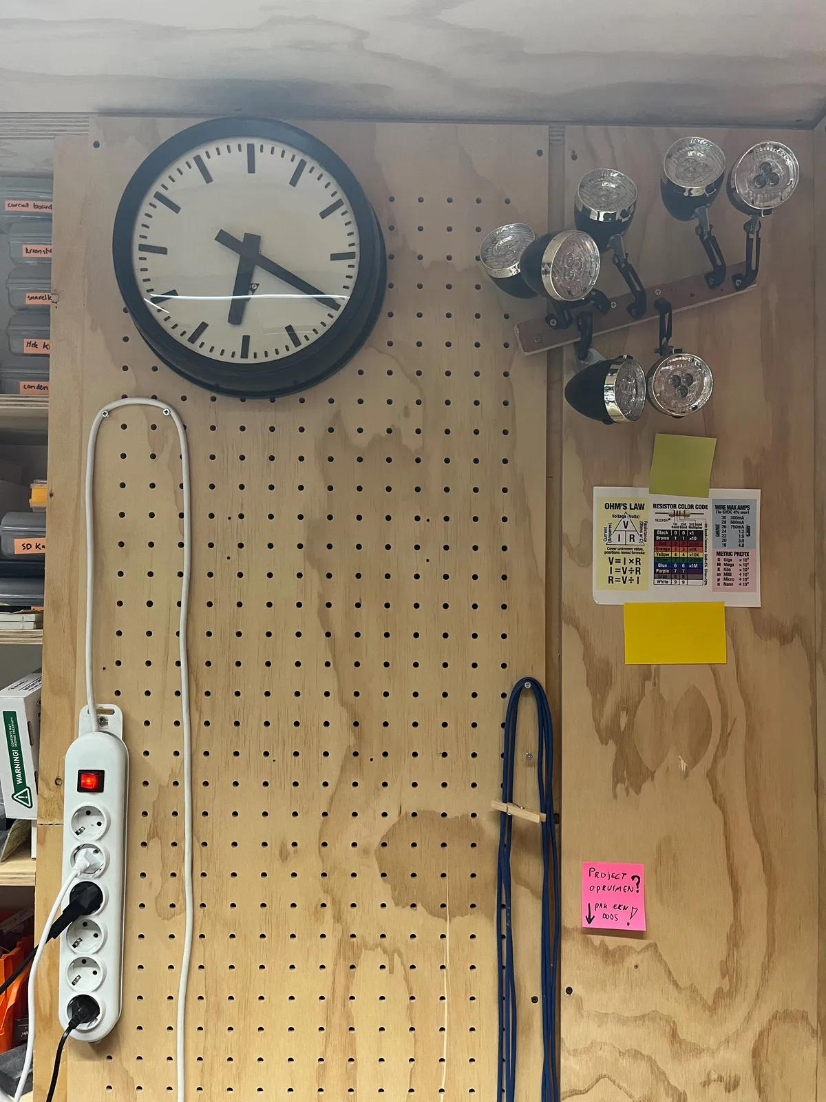 There are some plugs, some lamps, some wires and a clock in this part of a workshop. According to the clock it is 20 past six.