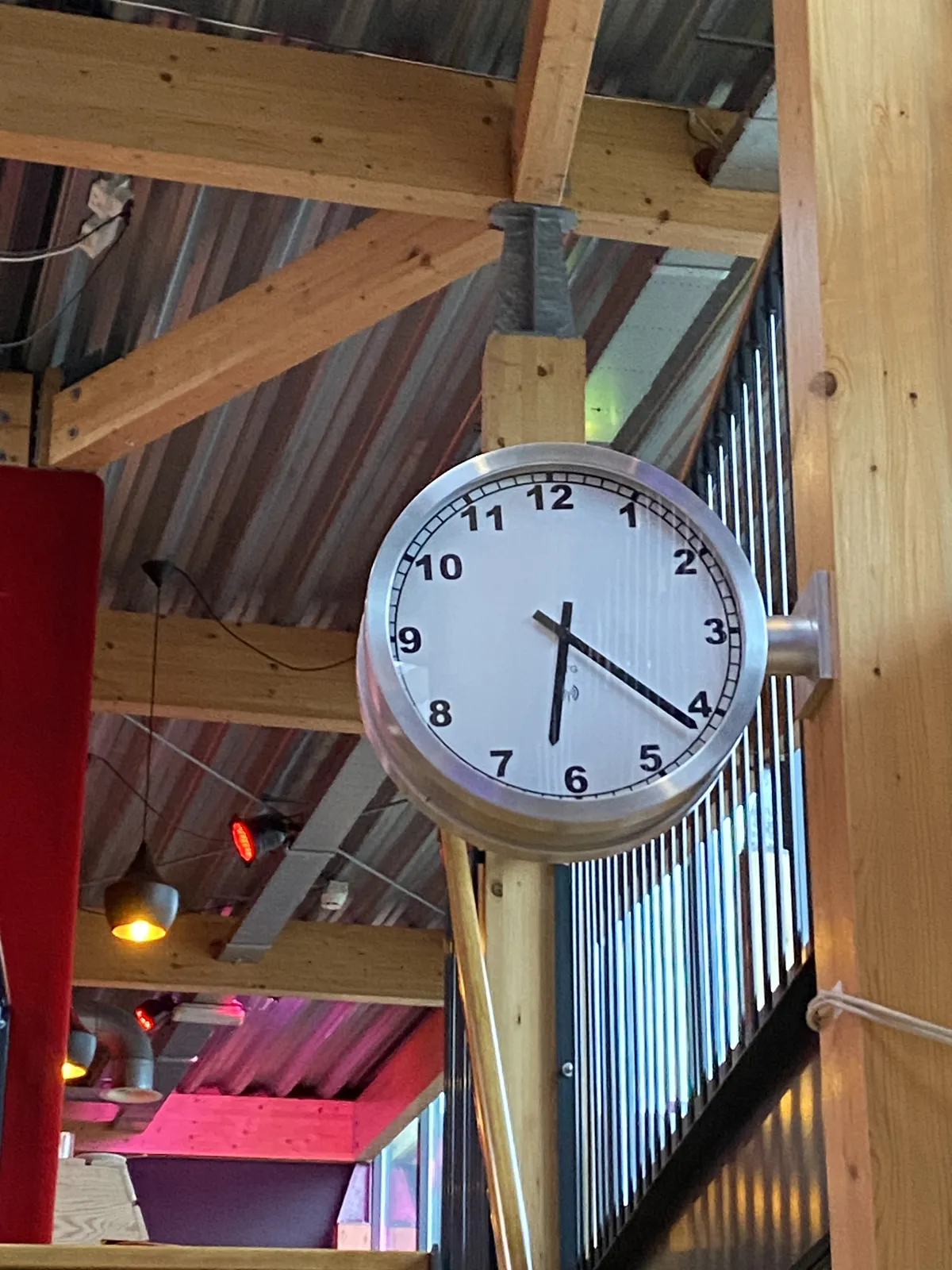 A large clock attached to a wooden beam in what seems like a modern cafe. It points at 21 minutes past 6.