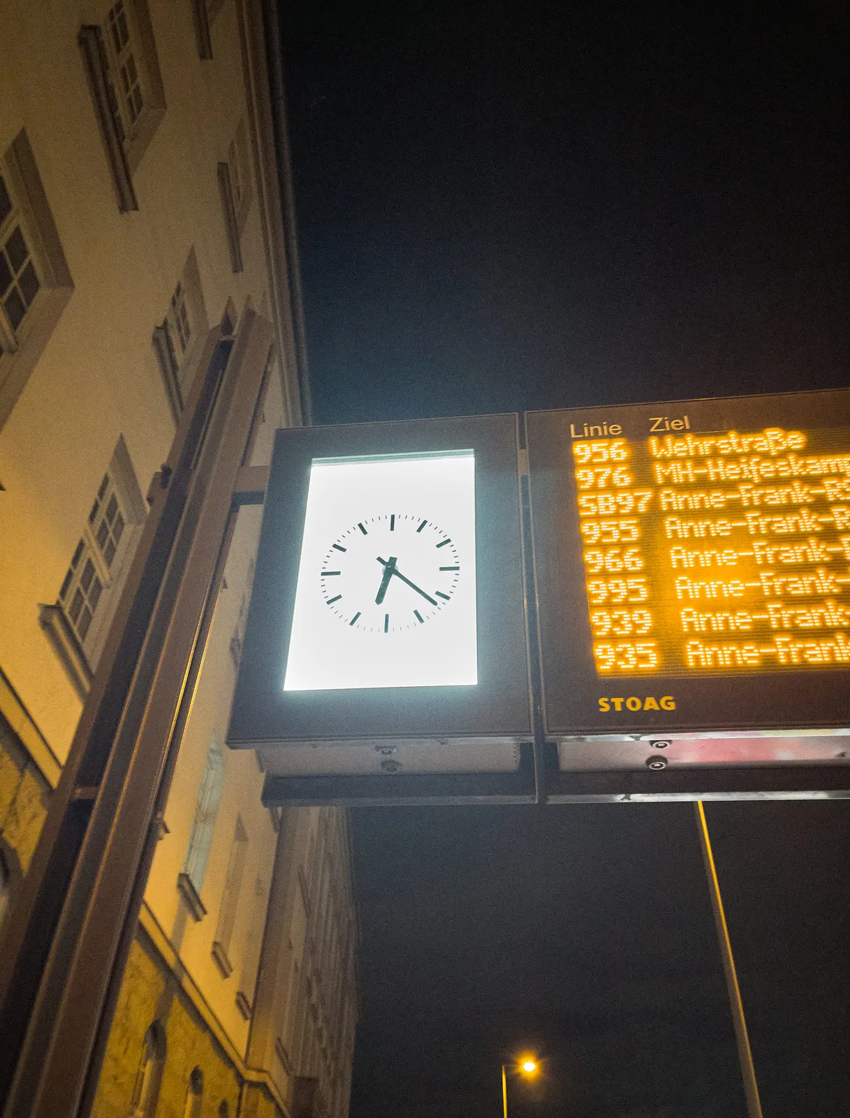 An appartment building in the back, a lit digital display with information about buses in the foreground. Most buses go to Anne Frank street. The clock next to the display points at 22 minutes past 6.