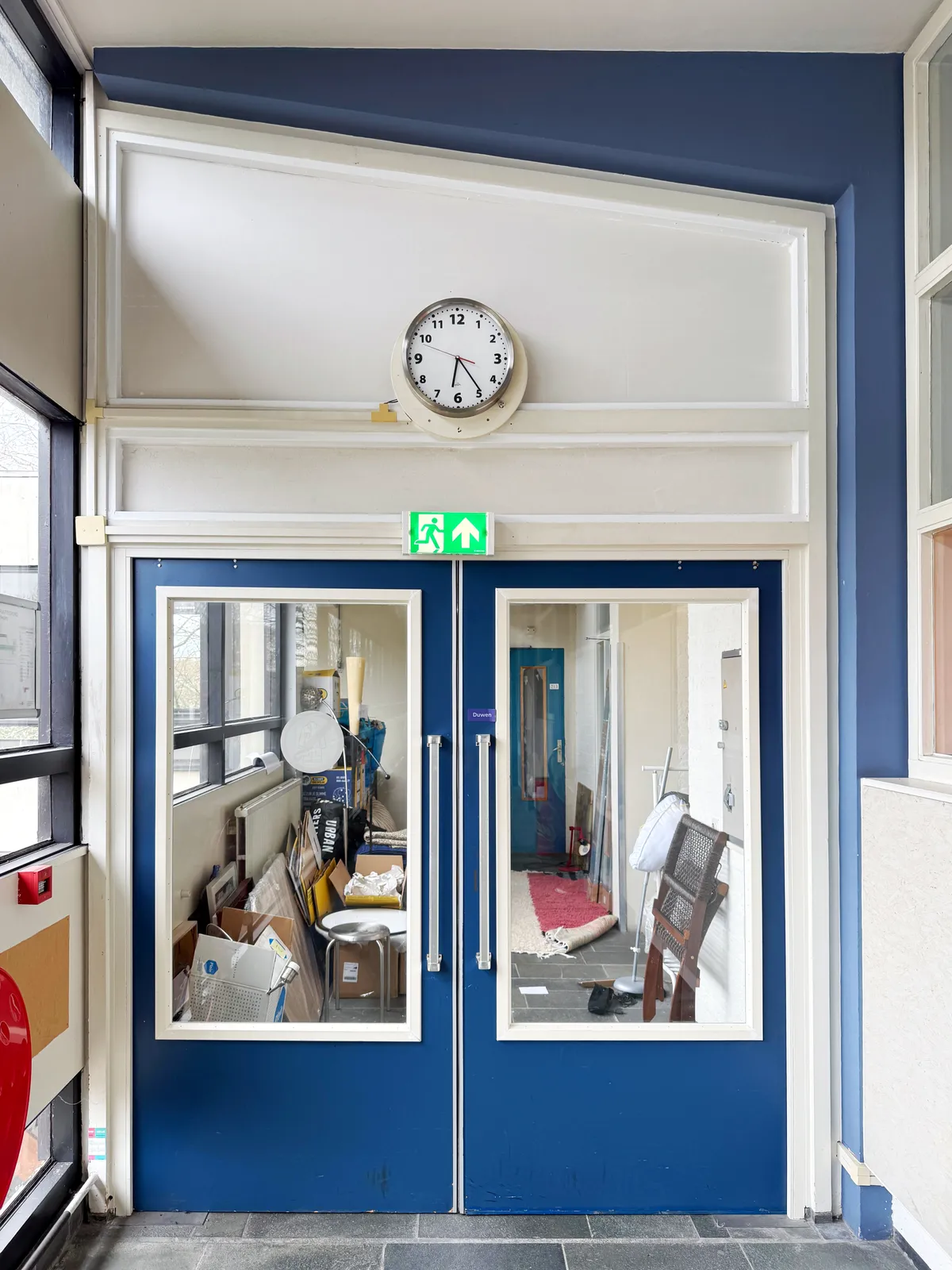 A wall clock above a double door. Behind the door there is a pile of stuff, as if someone is moving. The clock above the door points at 24 minutes past six.
