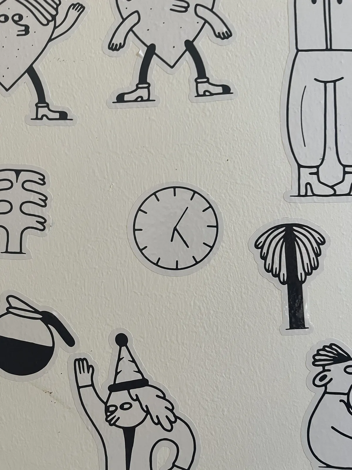 All kinds of happy stickers on a white wall. One of the stickers is a clock that points at 25 minutes past six.