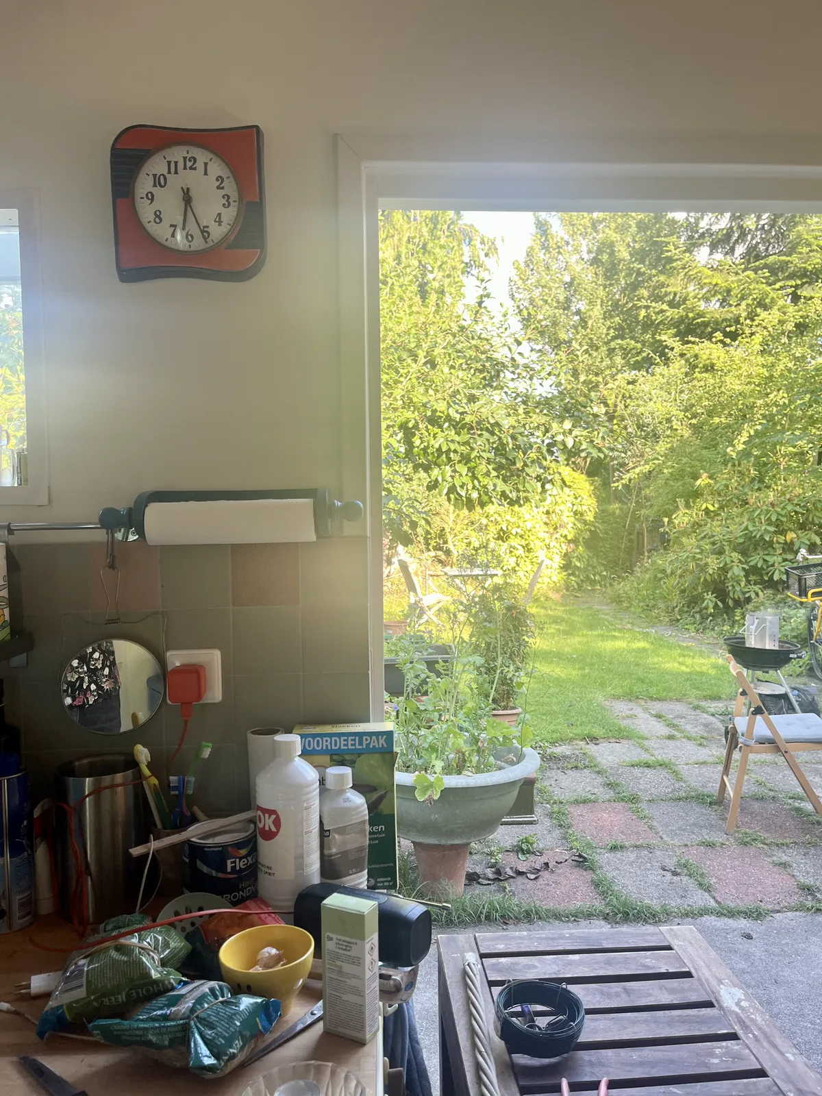 Standing in a kitchen, looking out through the open doors, into a very green garden. There’s an orange with black, ceramic clock on the wall in this kitchen. It points at 26 minutes past six.