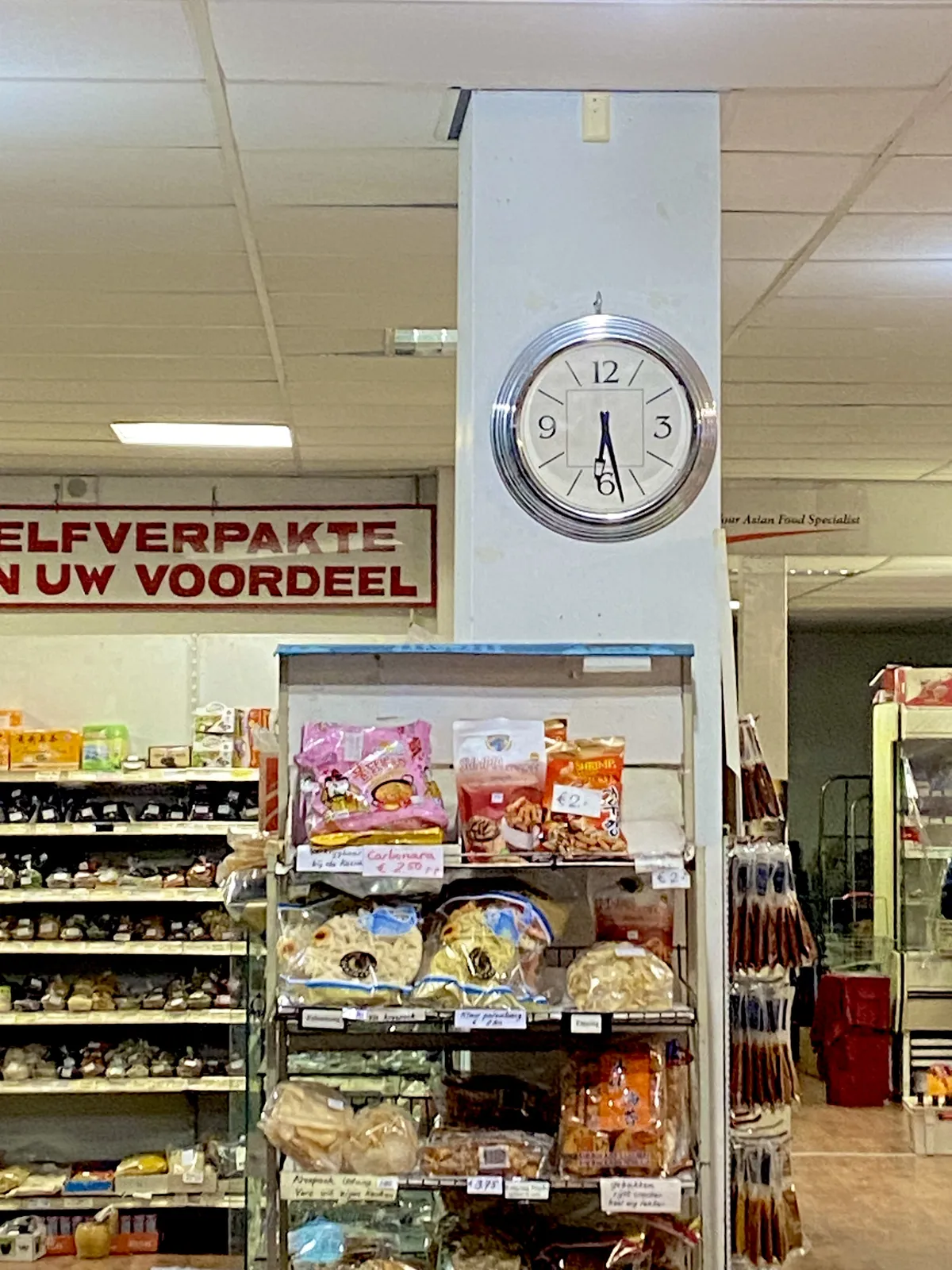 A clock on a pillar in a super market. It points at 28 minutes past 6.