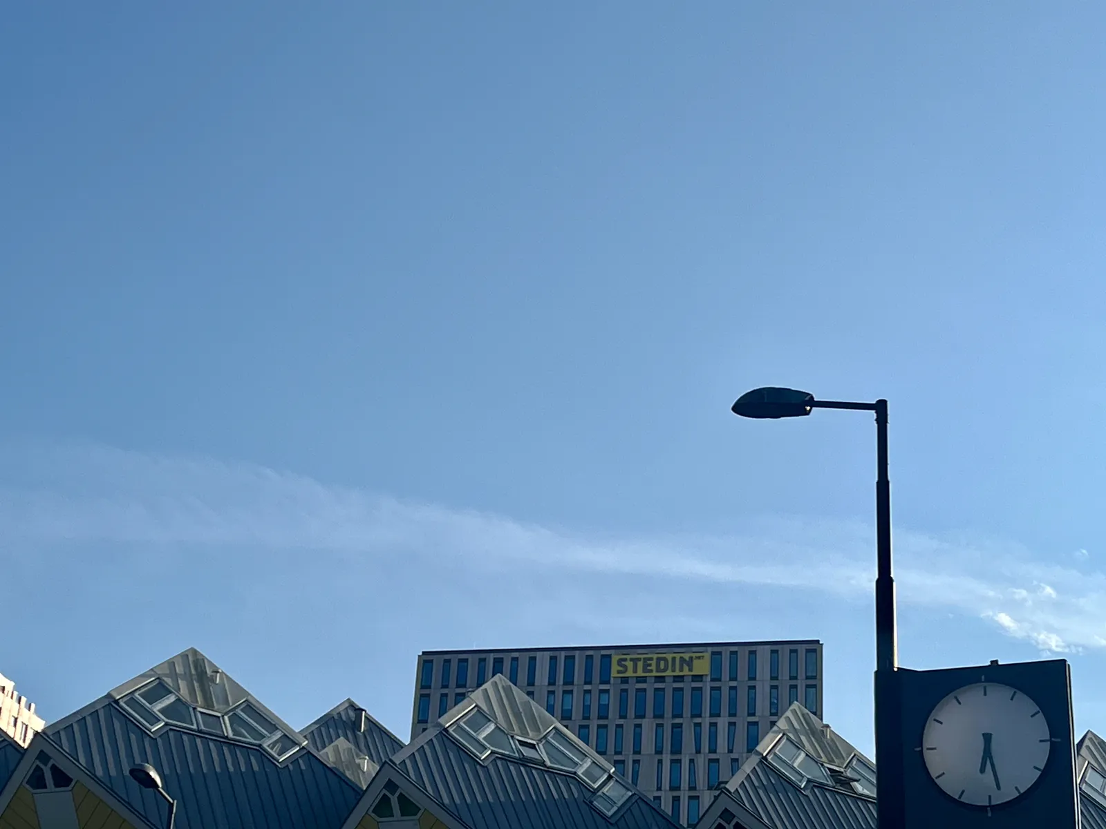 A clock attached to a pole. In the background you can see a bright blue sky, and the famous cube buildings of Rotterdam. On the clock it is 28 minutes past 6.