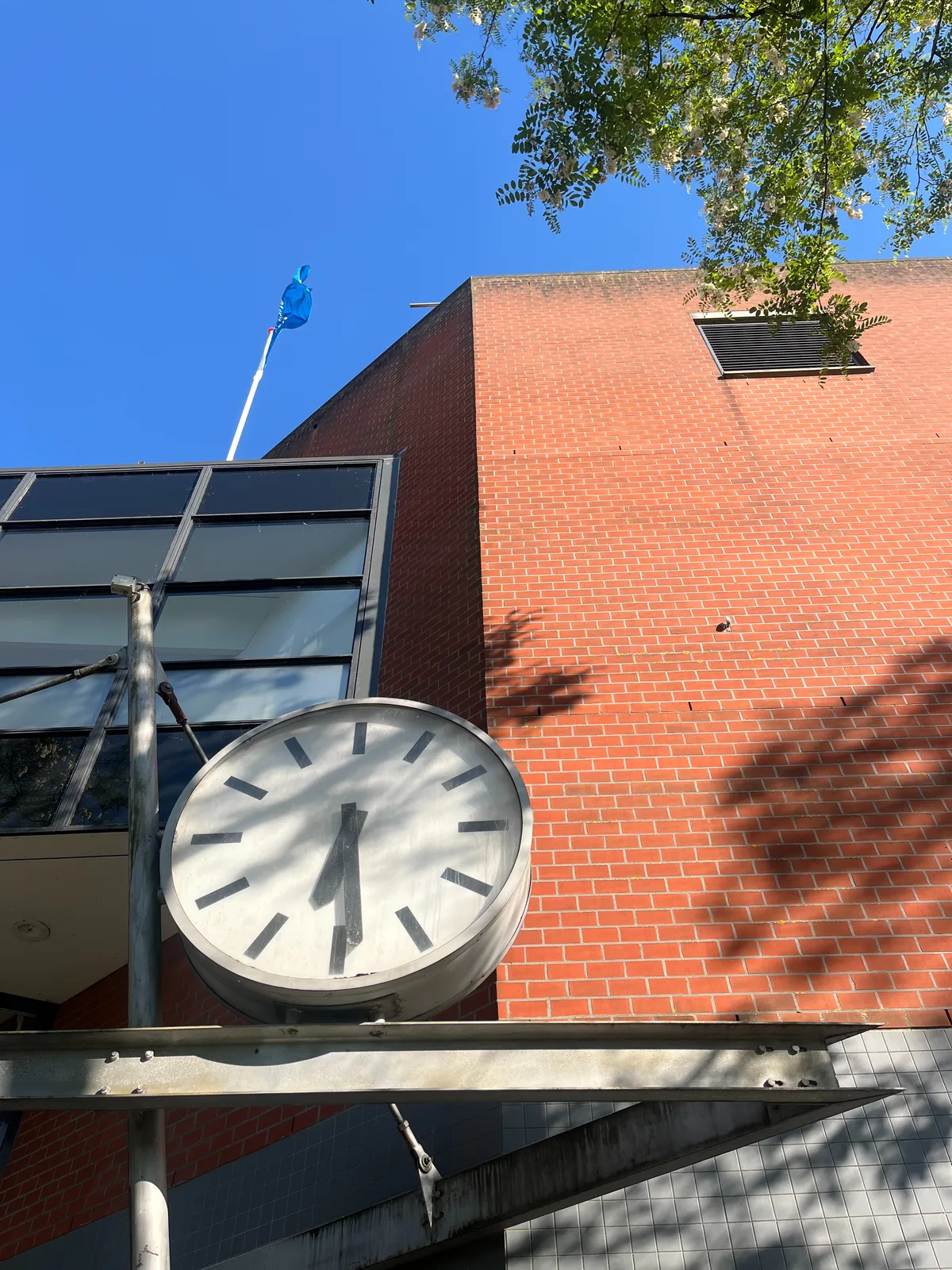 Looking up at the facade of a brick building, with some unclear constructions attached to it. There’s a large clock on this construction which points at 29 minutes past six