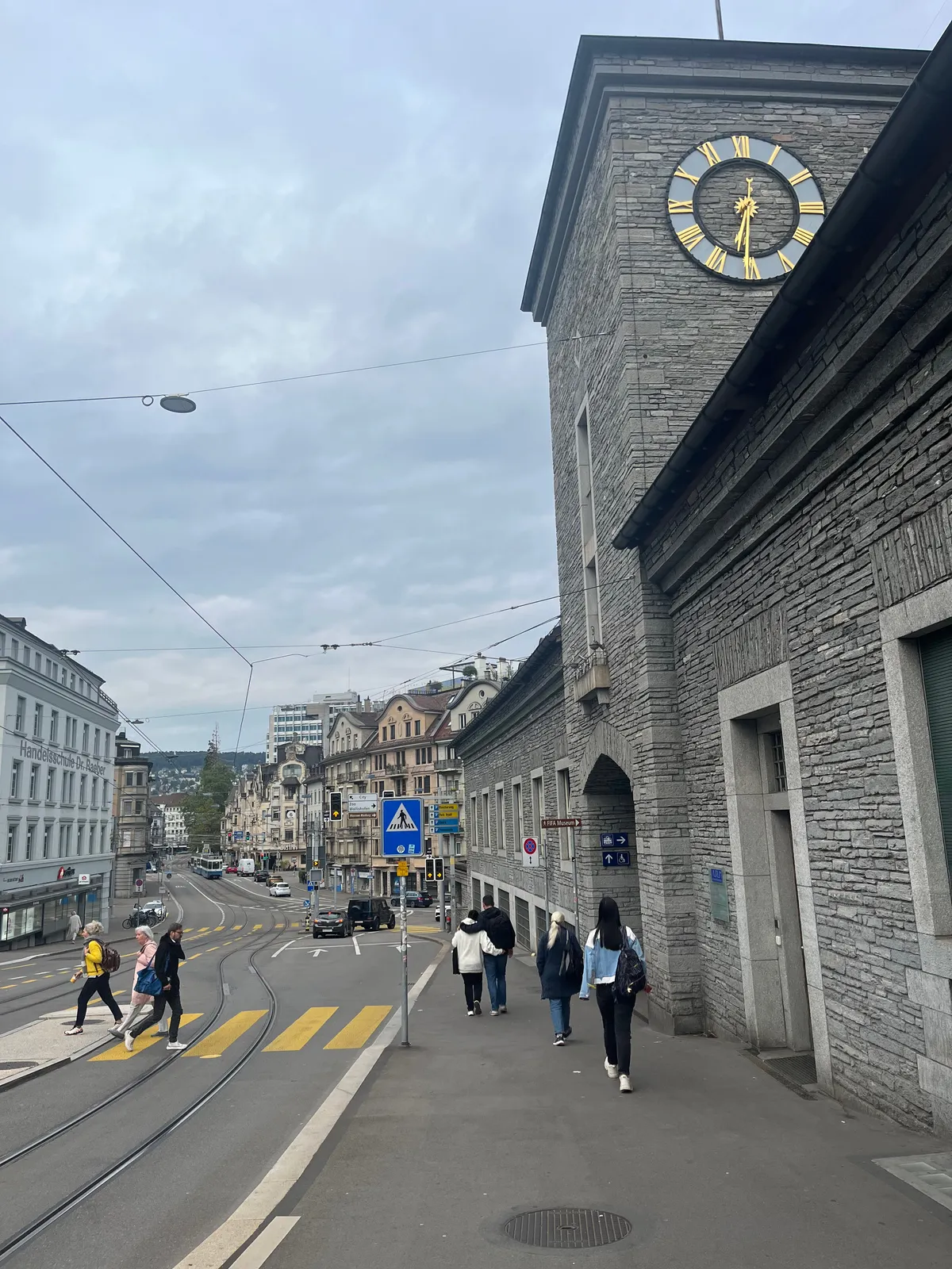 People walking on a hilly street in Zürich. On a solid train station building there’s a clock. On it it is 31 minutes past 6.