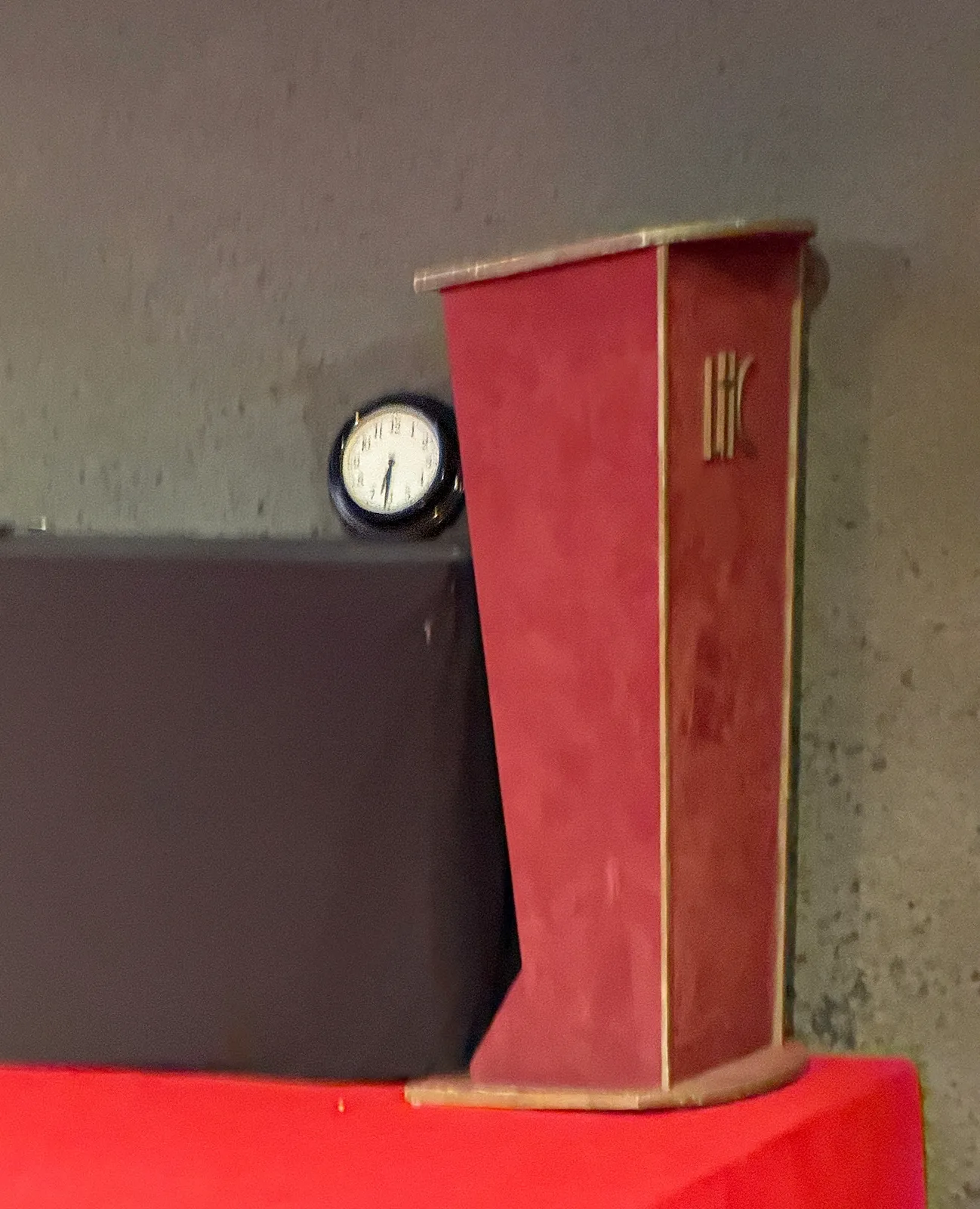 Next to a lectern, on a red carpeted stage there’s a wall clock that points at 31 minutes past six.