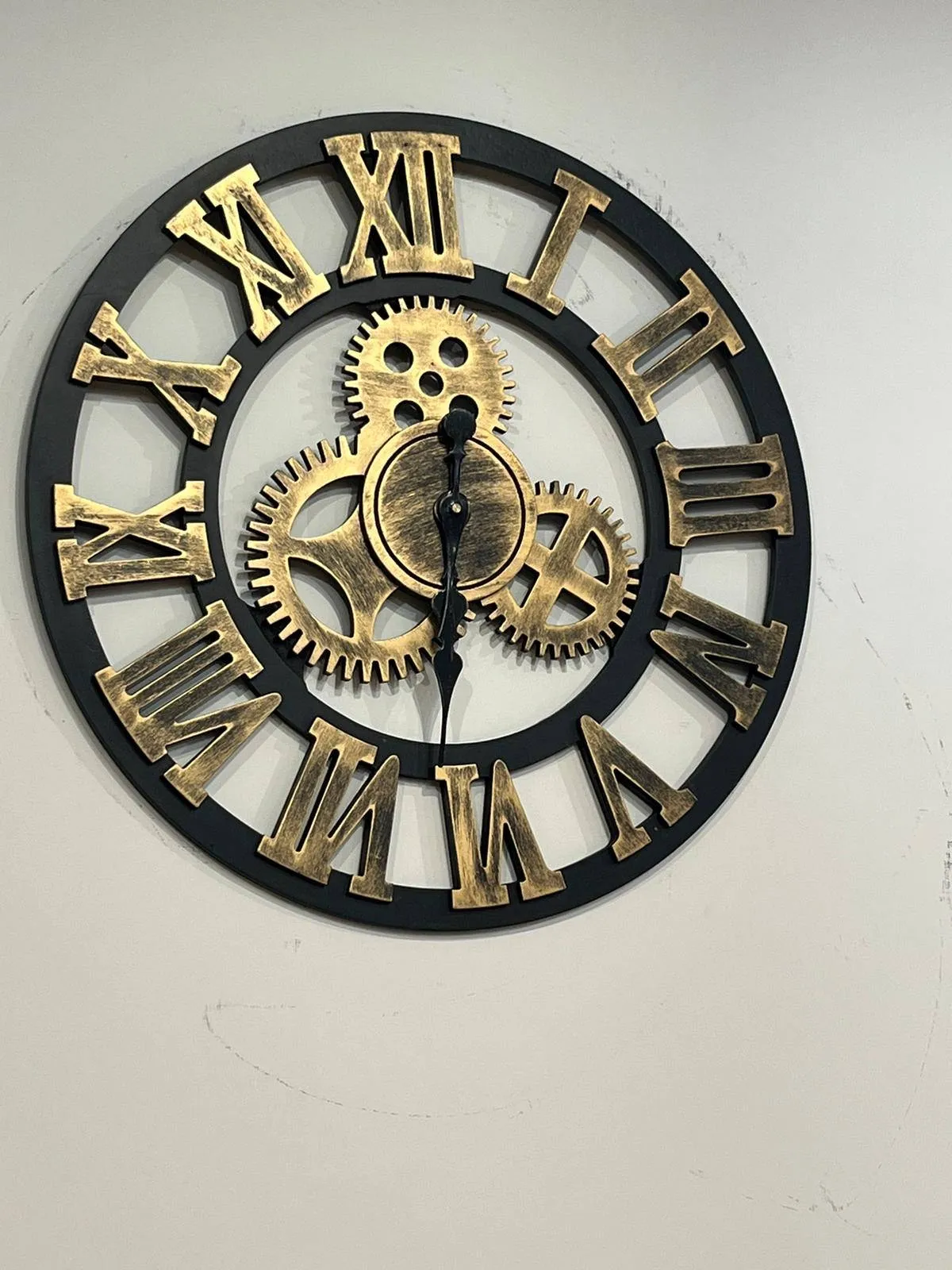 A clock made of two black circles with gold coloured, large roman numerals on it. In the center ring there are three large, gold coloured cogs that suggest that these are actual mechanics, the suggestion is not very strong though. The black classic hands both point down at approximately 28 minutes to 7.