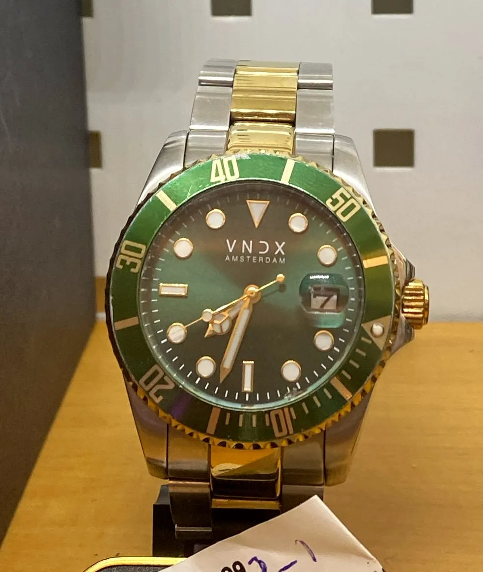 A green VNDX Amsterdam wrist watch. It points at 27 minutes to 7.