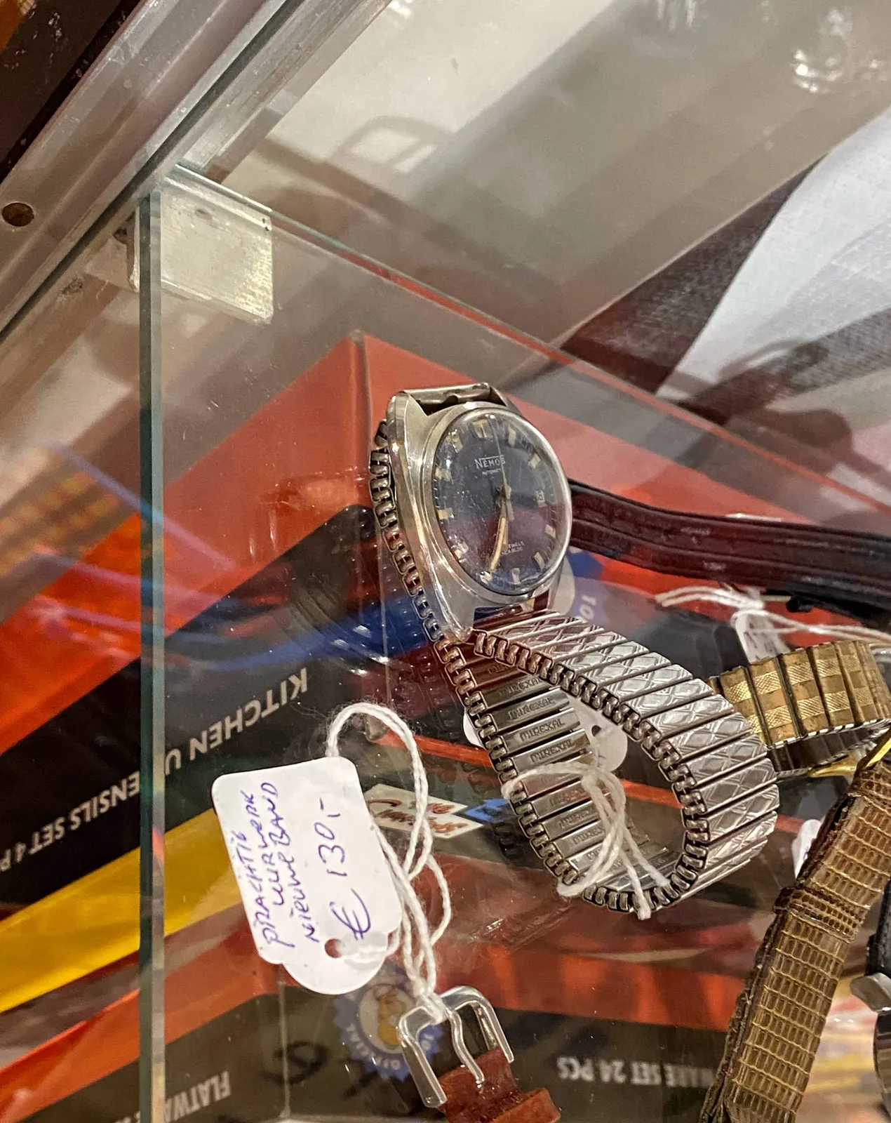 A wrist watch with a silver strap on a glass plate, it’s for sale for 130 euros. Below it are some books. The watch points at 26 minutes to 7.