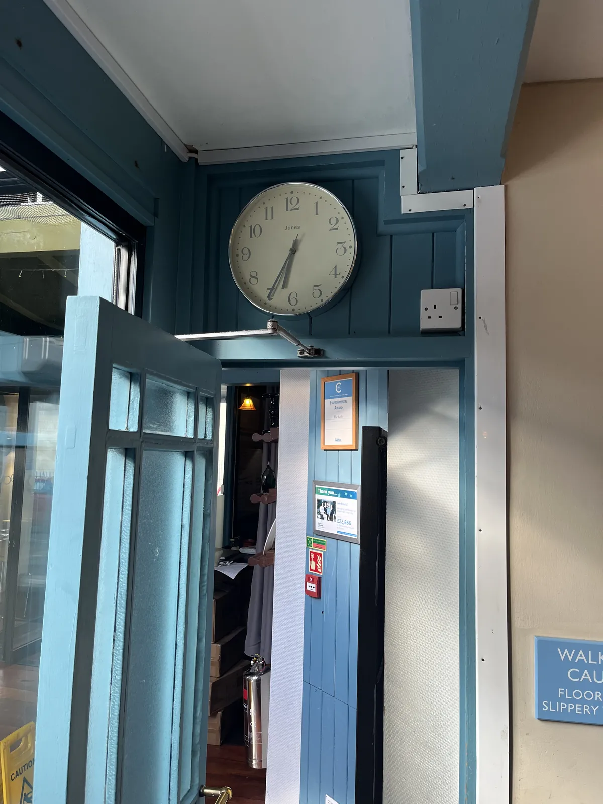 An open wooden door, wooden frames, some signs, and mostly blue. The clock above the door points at 35 minutes past six.