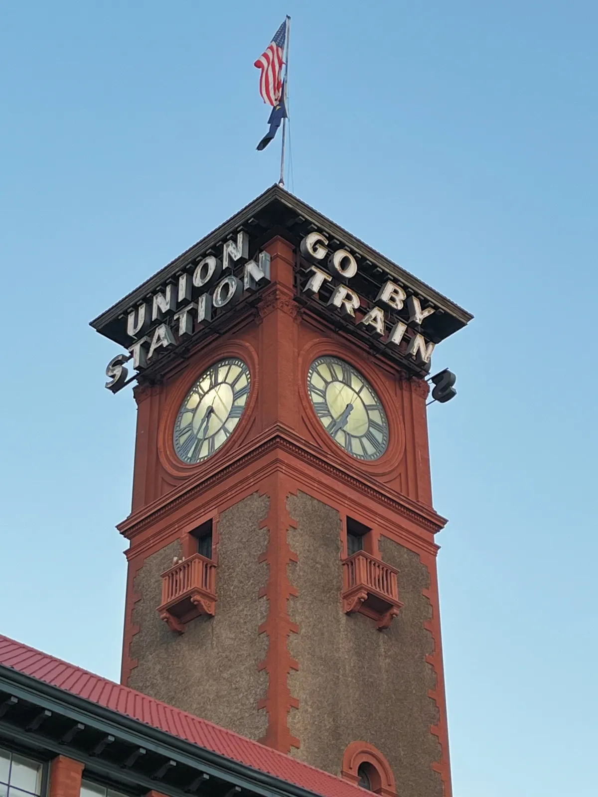 A red roof with a mostly red tower on it with a US flag op top, and a message that says this is a Union station and which urges you to go by train. The clock on this tower points at 25 minutes to 7.