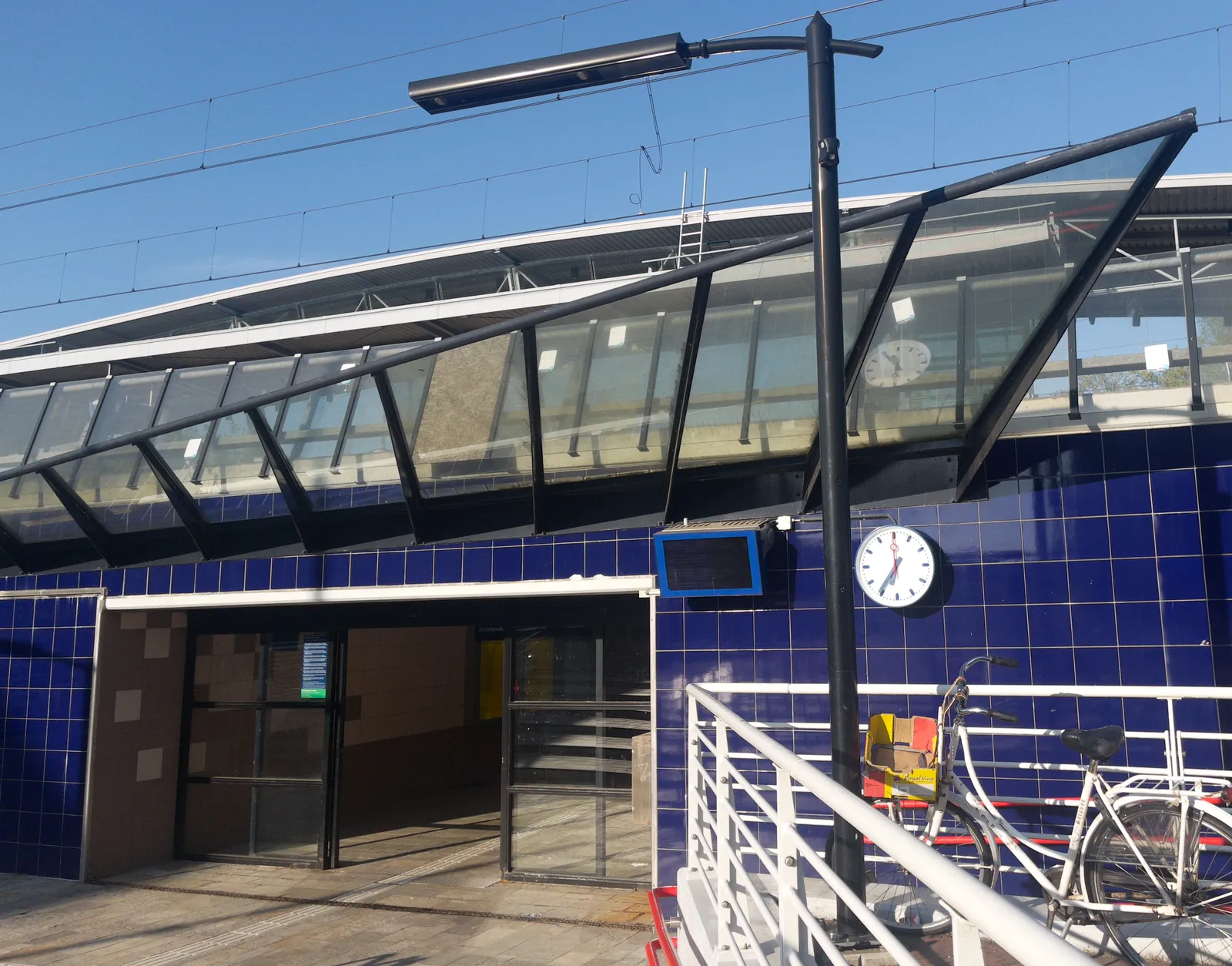 The entrance to a small Dutch railway station. It has dark blue tiles around the doors, a strange, random triangular glass structure above the doors, and a clock next to the door. This clock points at 24 minutes to 7.