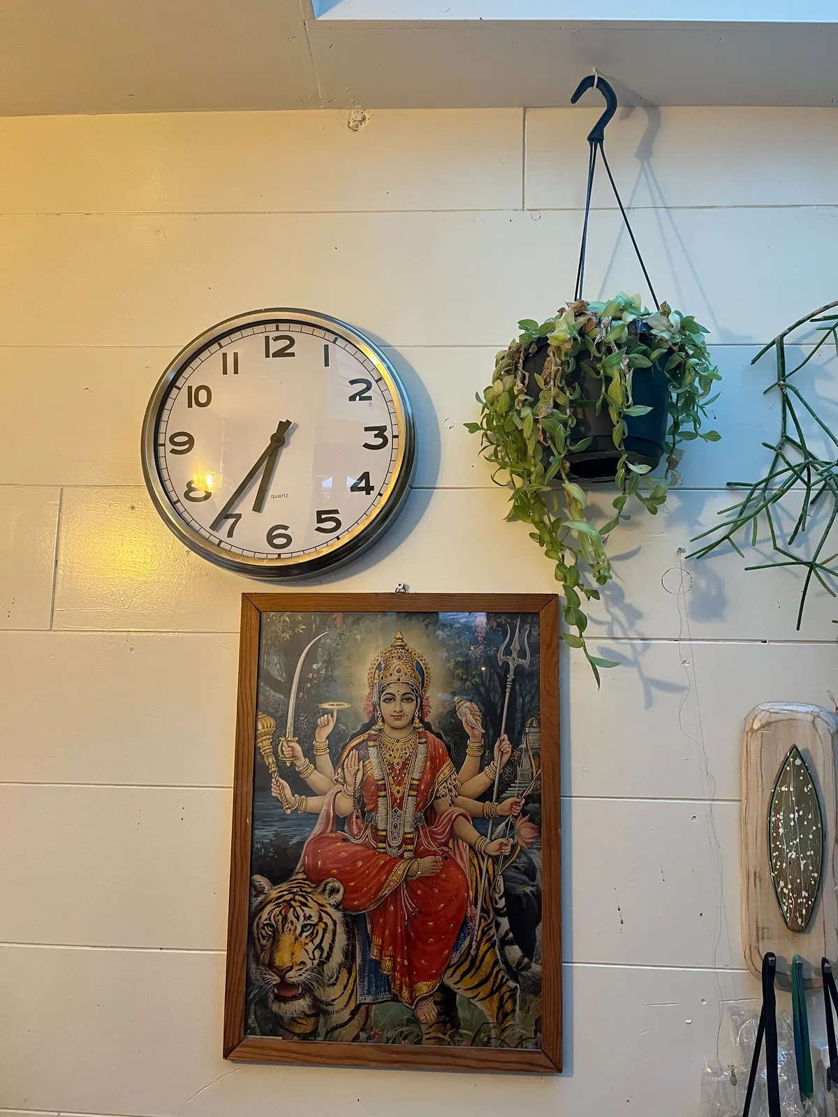 In this picture there's a plant hanging from the ceiling, a picture of a multi-armed goddess sitting on a lion, and a clock which points at 24 minutes to 7. 