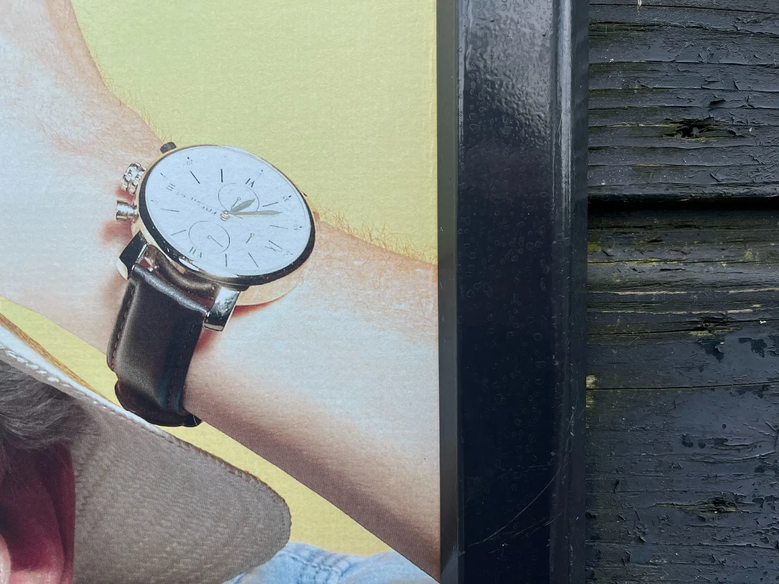 A closeup of someone’s wrist with a watch on it. On a closer look it turns out this is a detail of a bilboard on a fence. The watch points at 23 minutes to 7.