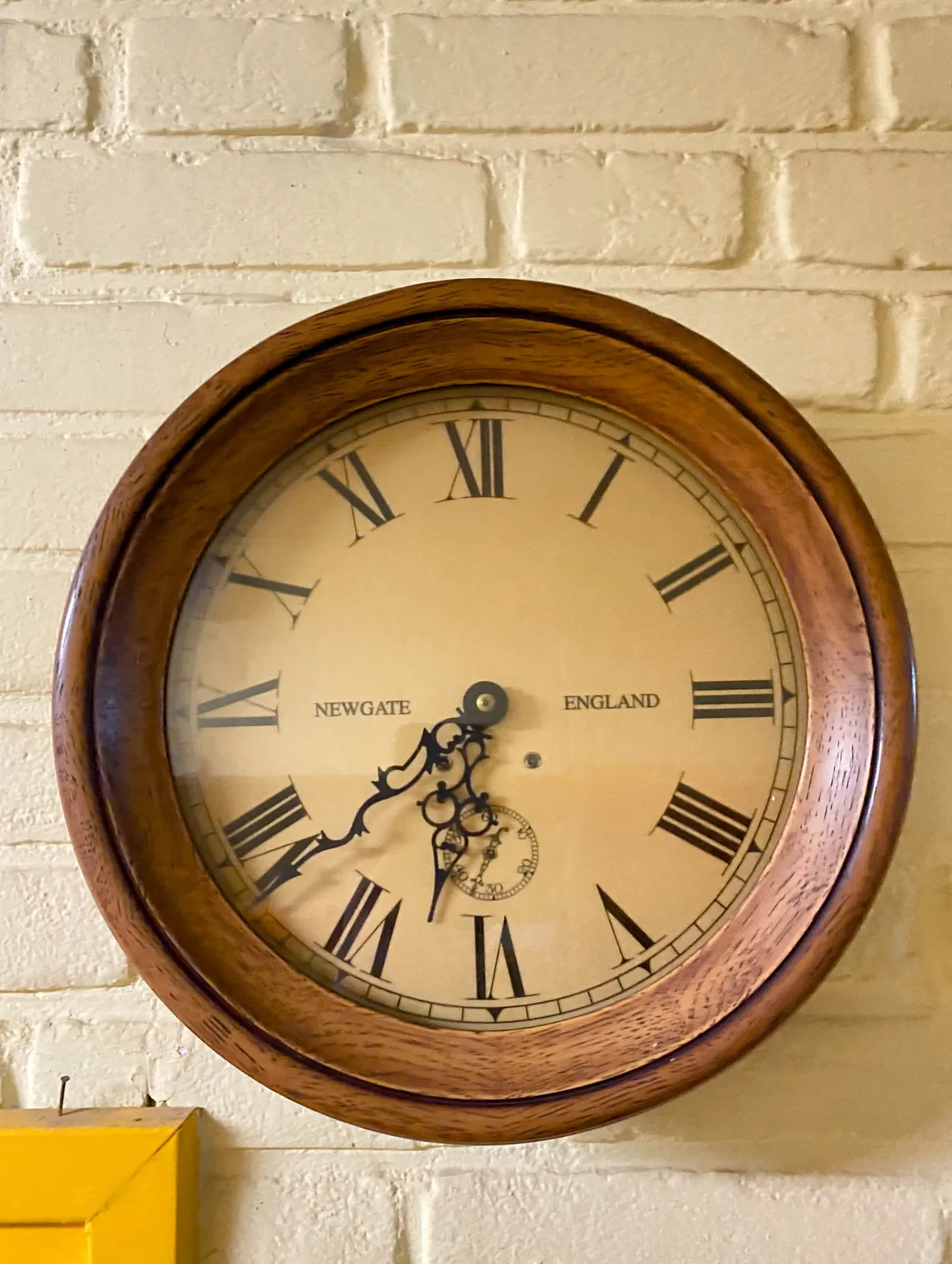 A round wall clock in a wooden frame. The words Newgate England and a fake dial are printed on its face. The clock points at 22 minutes to 7.