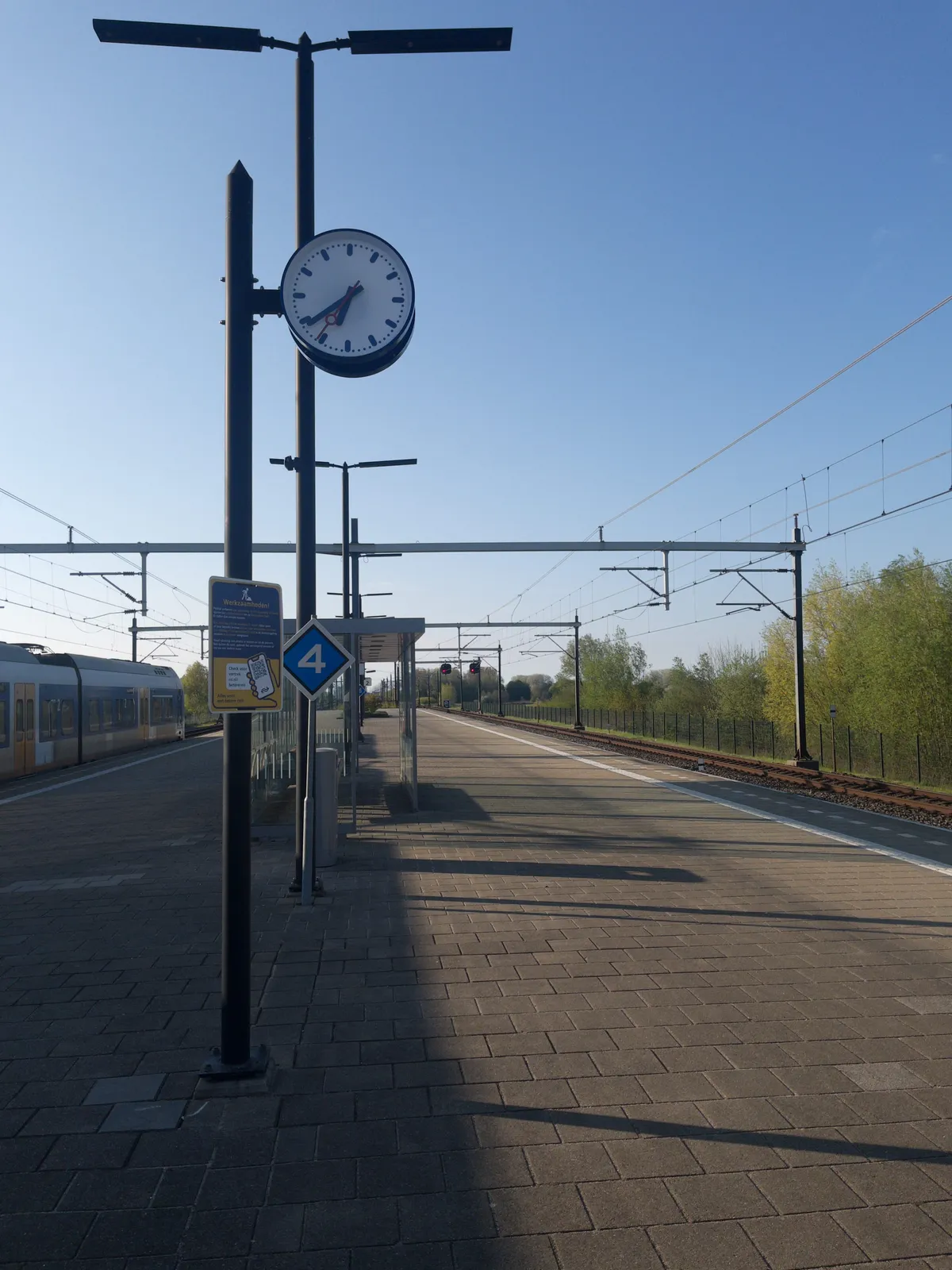 A clock on a pole on an empty platform of a rural Dutch train station. It points at 21 minutes to 7.