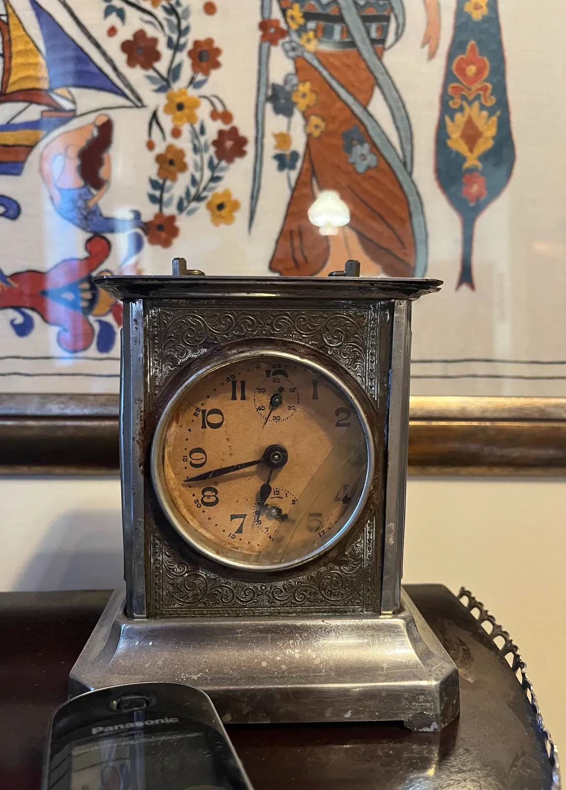 An old, silver clock, with a framed piece of embroidery in the background. On the clock it is 18 minutes to 7