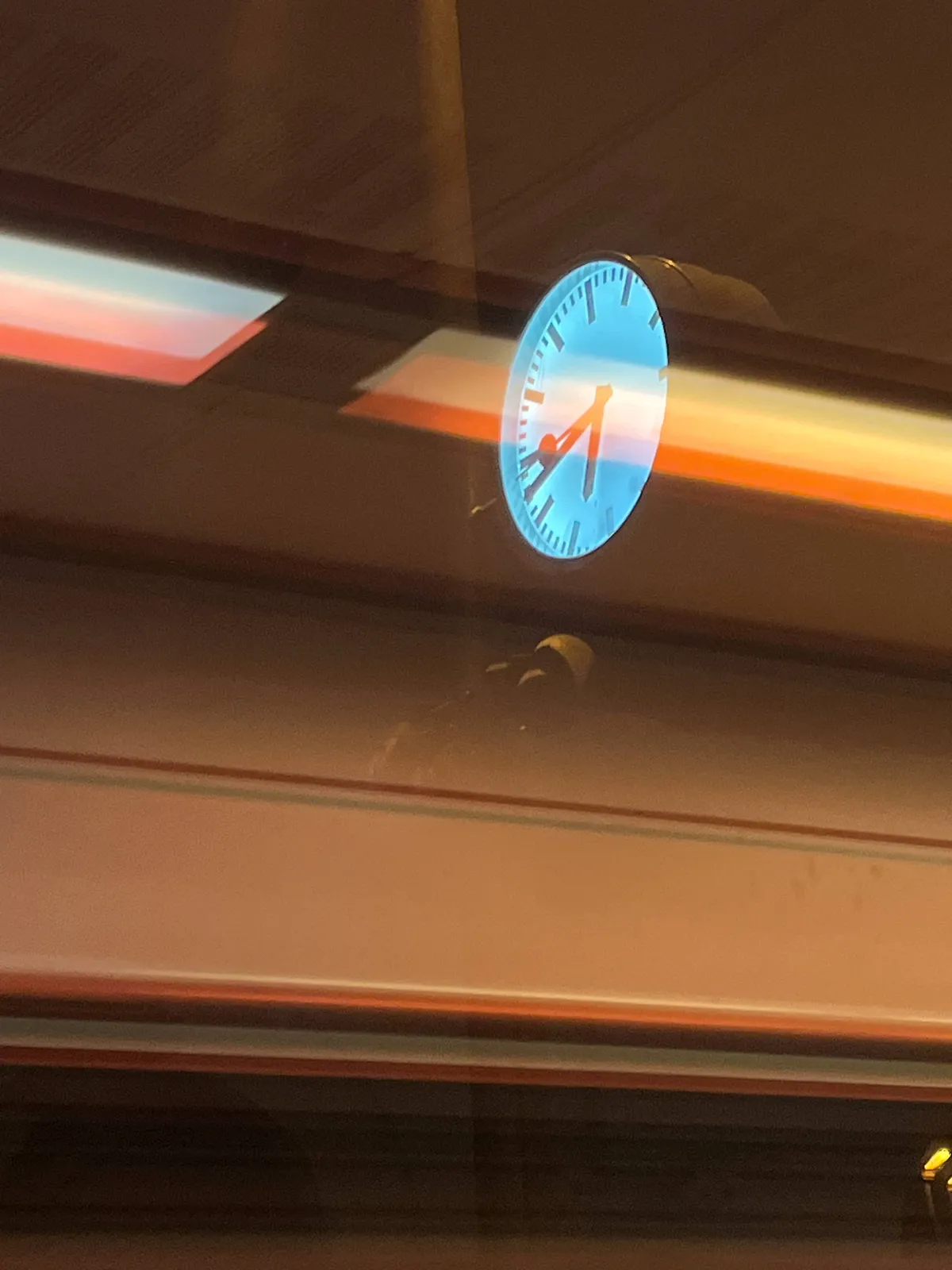 Looking out through a window which is reflecting lights. Outside there’s a lit clock. It points at 42 minutes past six. 