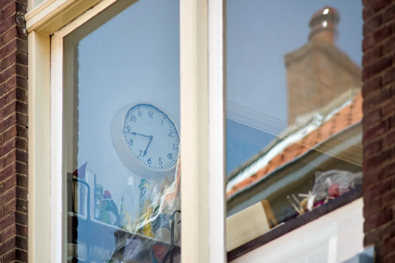 Looking through an old window. Behind the window there’s a clock that looks distorted. The effect is caused by the wobbly old glass. The clock points at 17 minutes to 7. 