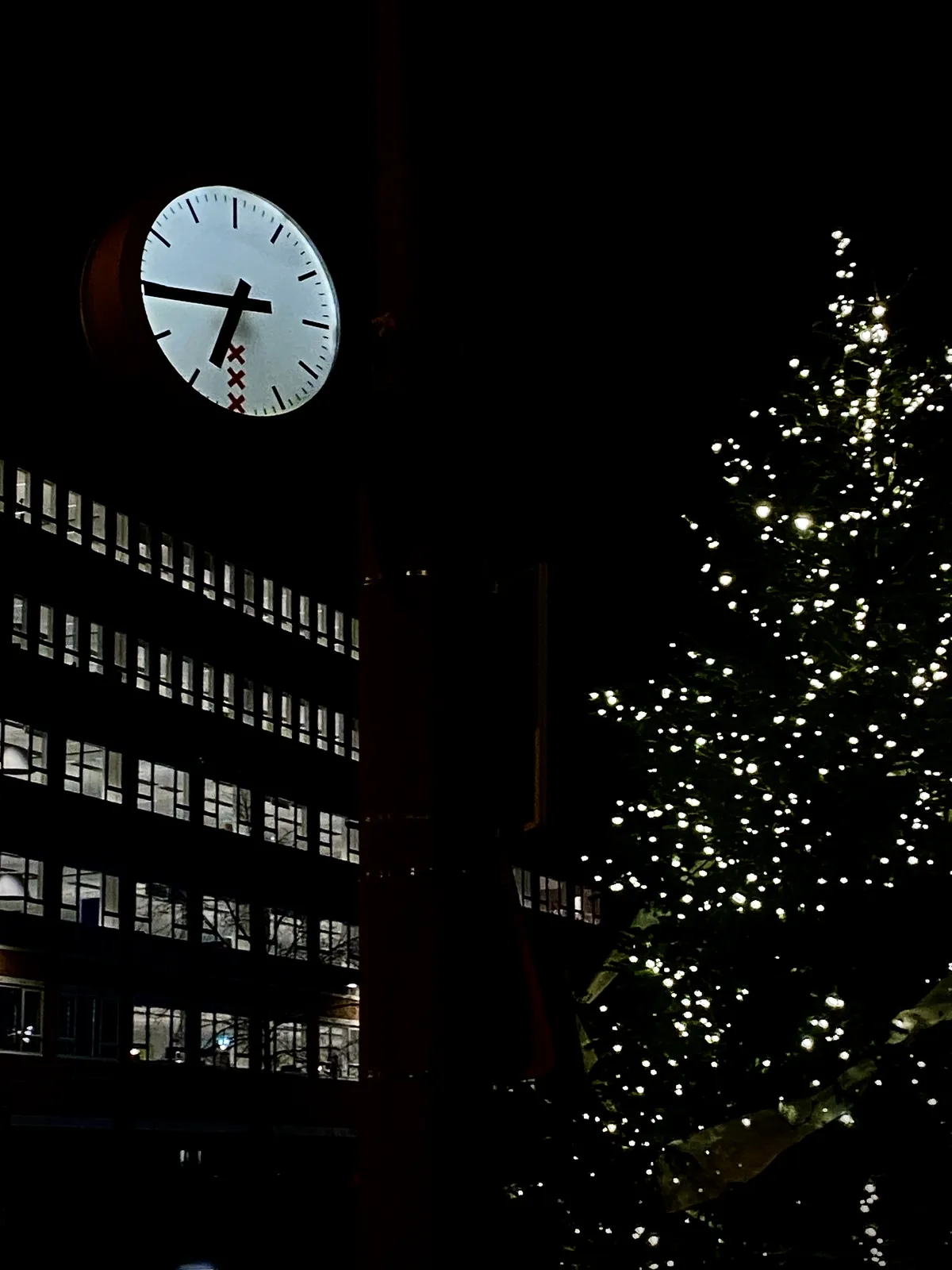 It’s very dark outside. All the windows in a large building in the background are lit. There’s also a christmas tree with lights in it. And there’s a lit clock with the logo of Amsterdam in it. It points at 16 minutes to 7.