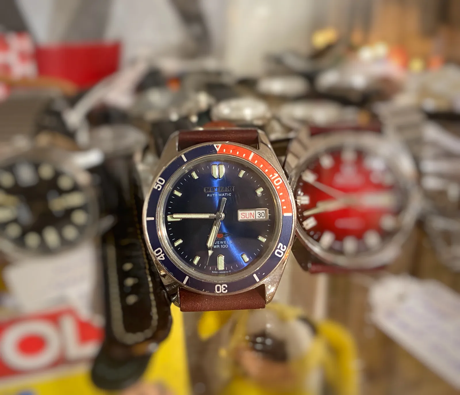 Blurry watches in the background, one watch in focus in front. It points at a quarter to 7.
