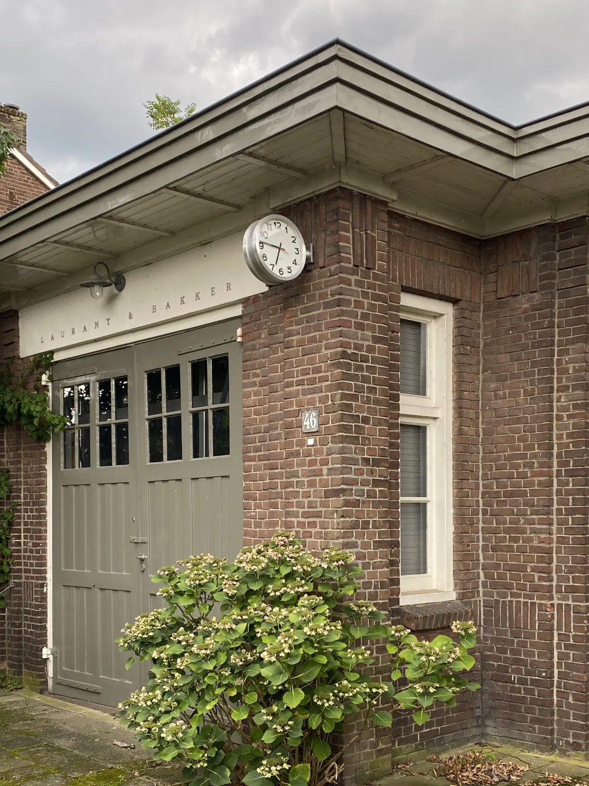 A brick building with wooden doors. Above the doors it says Laurent &amp; Bakker. There’s a clock attached to the building. It points at 14 minutes to 7.