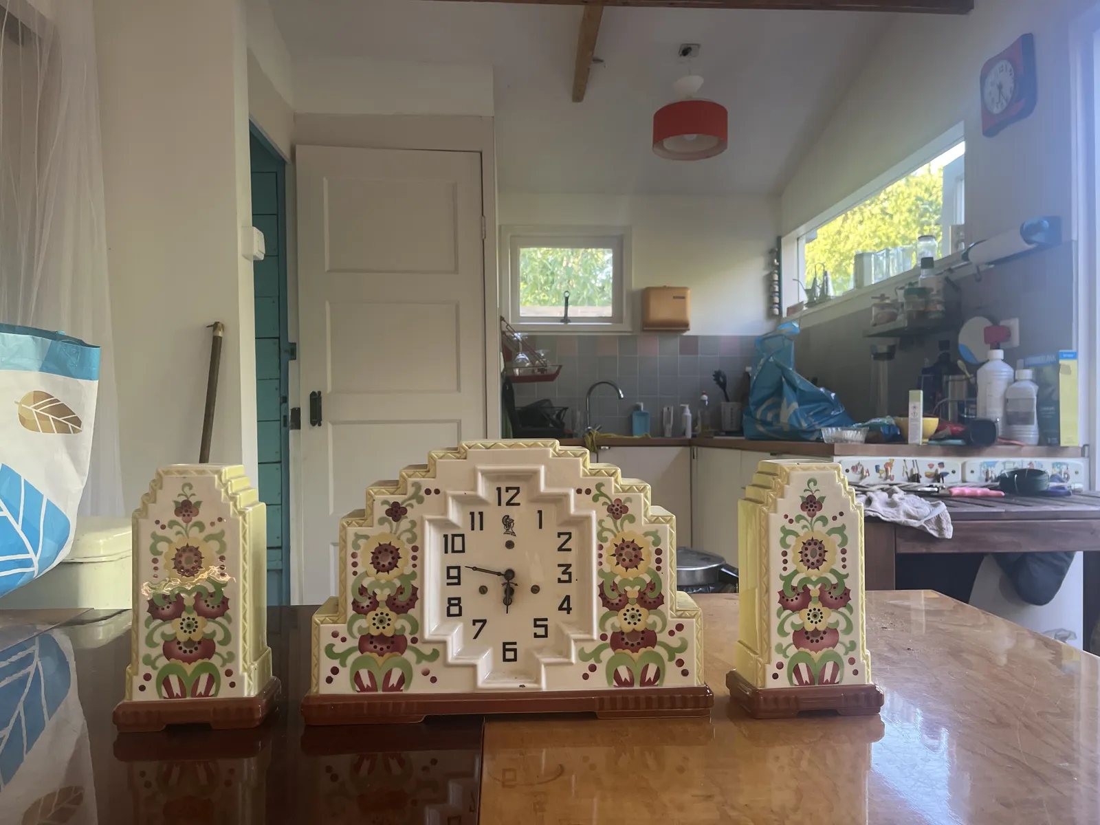 A ceramic mantel clock, with matching tulip vases, standing on a table. It has flowers painted on it, and the shape is geometric. It points at 13 minutes to seven. 