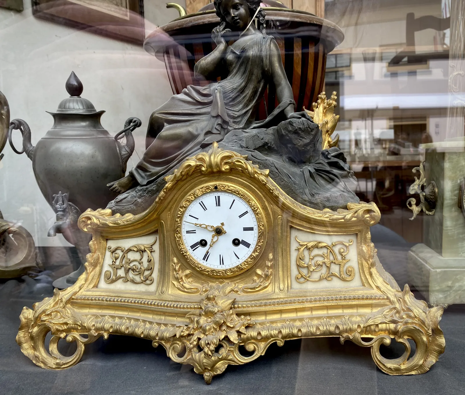 A golden, richly ornamented mantel clock, with a black statue of a sitting woman on top. The clock points at 12 minutes to 7.