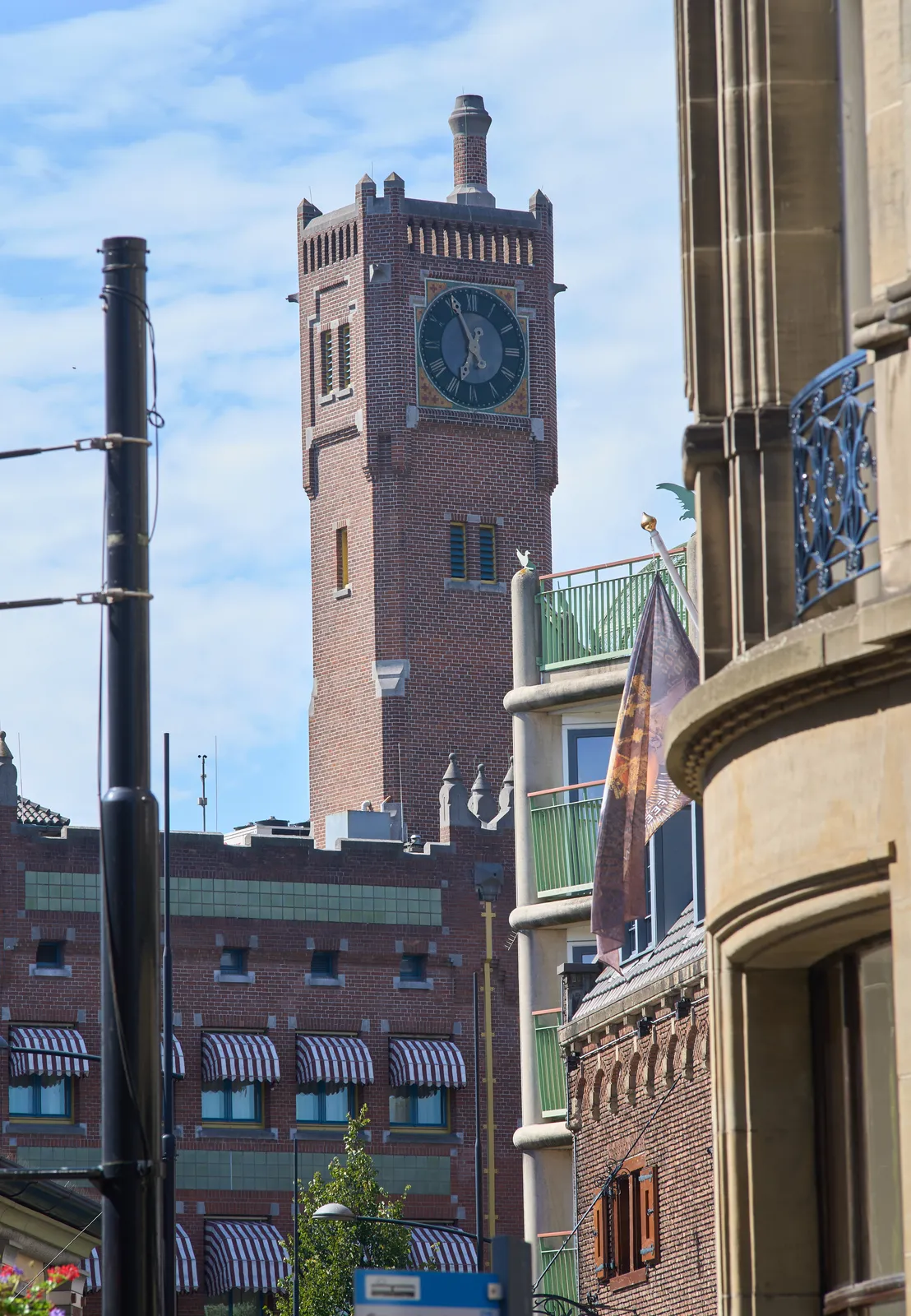 On the right, in the foreground, an old building. On the left a black pole. In the background a castle-like building with a rather high tower with a clock on it. It points at 4 minutes to 7.