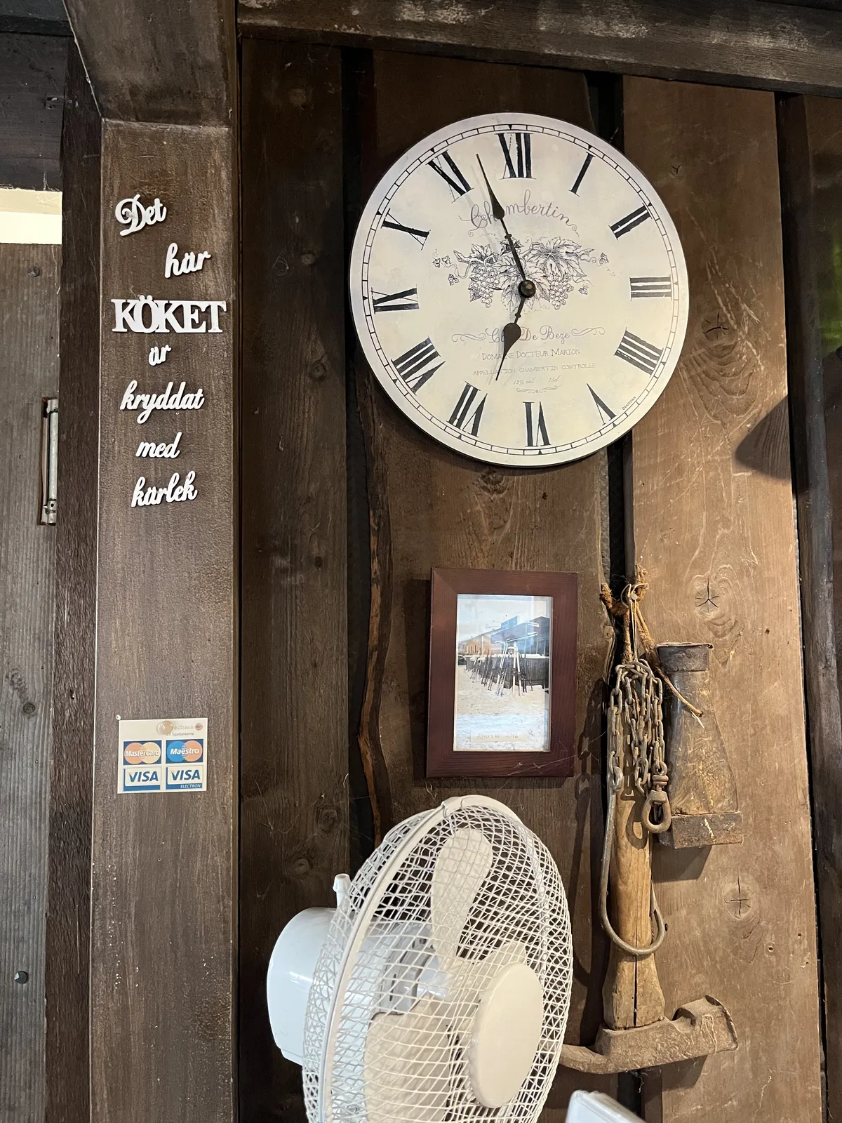 On the wall it says, in Swedish: This kitchen is seasoned with love. A ventilator stands on the table, and above it, also on the wall, there’s a clock that points at 3 minutes to seven