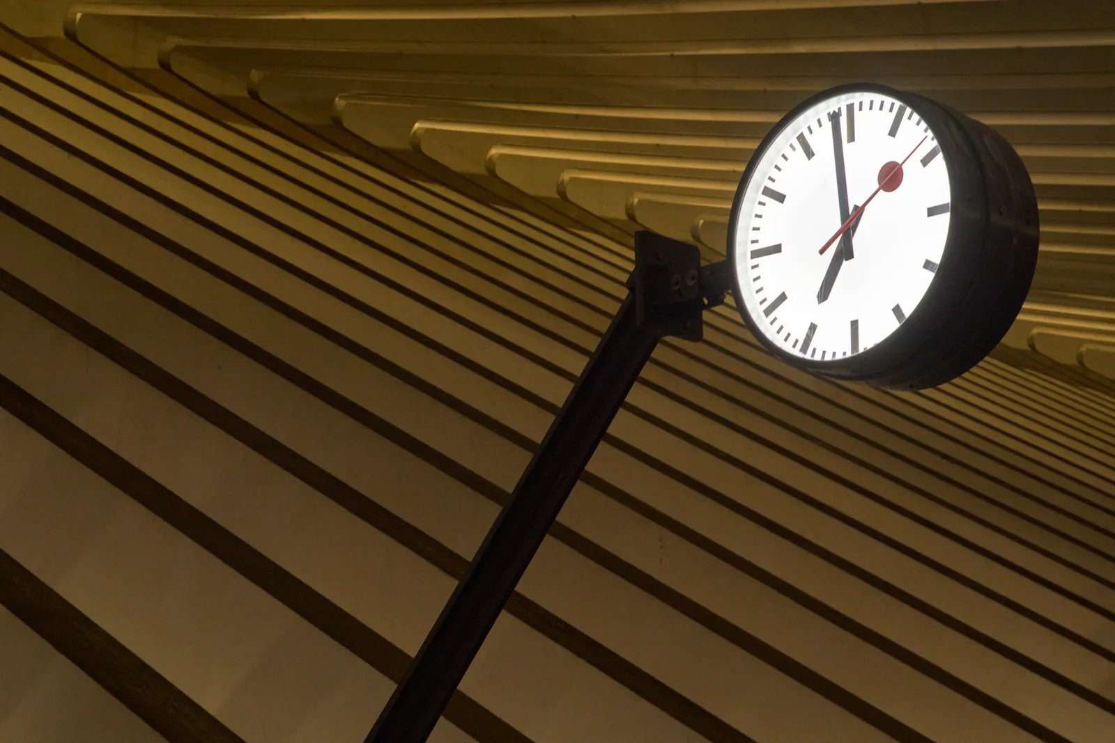 A station clock in front of a geometric, repeating construction. It points at 2 minutes to seven.
