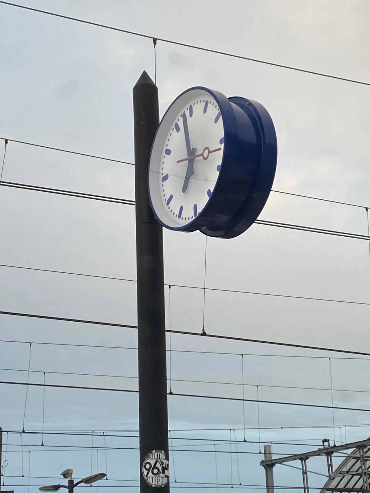 A clock on a pole. Probably on a train station, there are many wires behind it. It points at 2 minutes to 7.