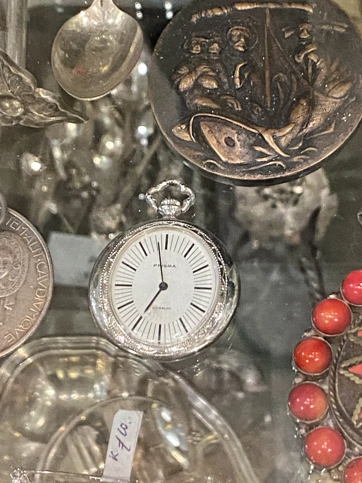 Some silver things on a glass plate. Among them a silver clock. It points at 2 minutes to 7.