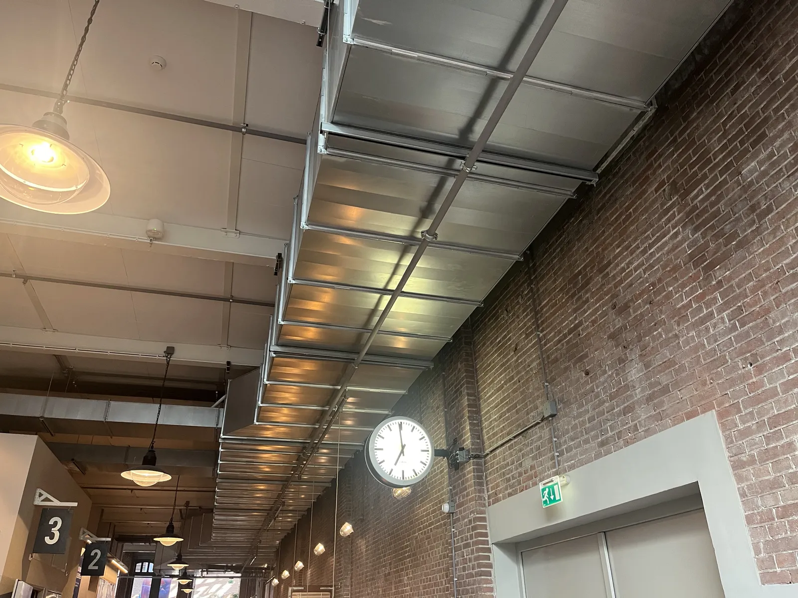 A clock attached to a long brick wall, below a large airconditioning structure, inside a building. The clock points at one minute to seven.