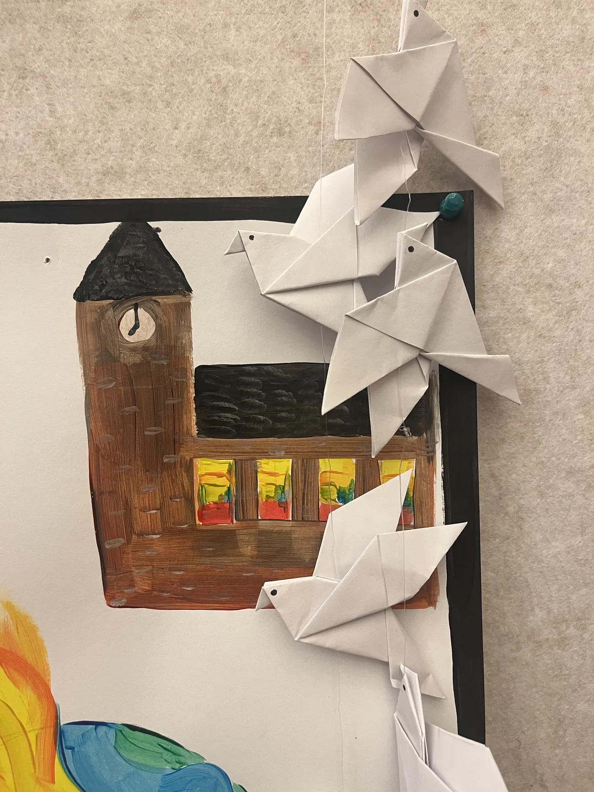 A childs painting of a church, with some origami birds in front of it. There’s a clock painted on this church. It is a bit unclear what time it is: the minute hand points straigh up, but the hour hand is right between seven and eight. So it’s either seven or eight o’clock.