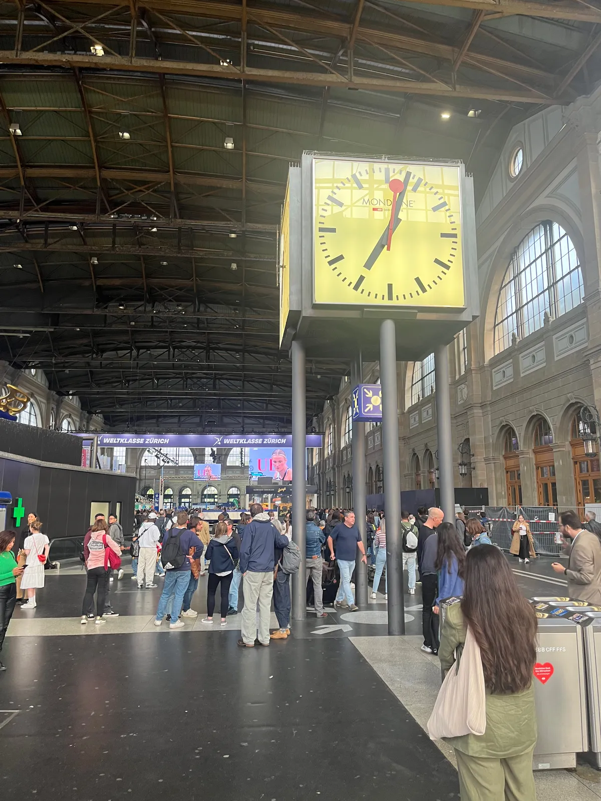 An oversized clock in a crowded train station points at 3 minutes past 7.