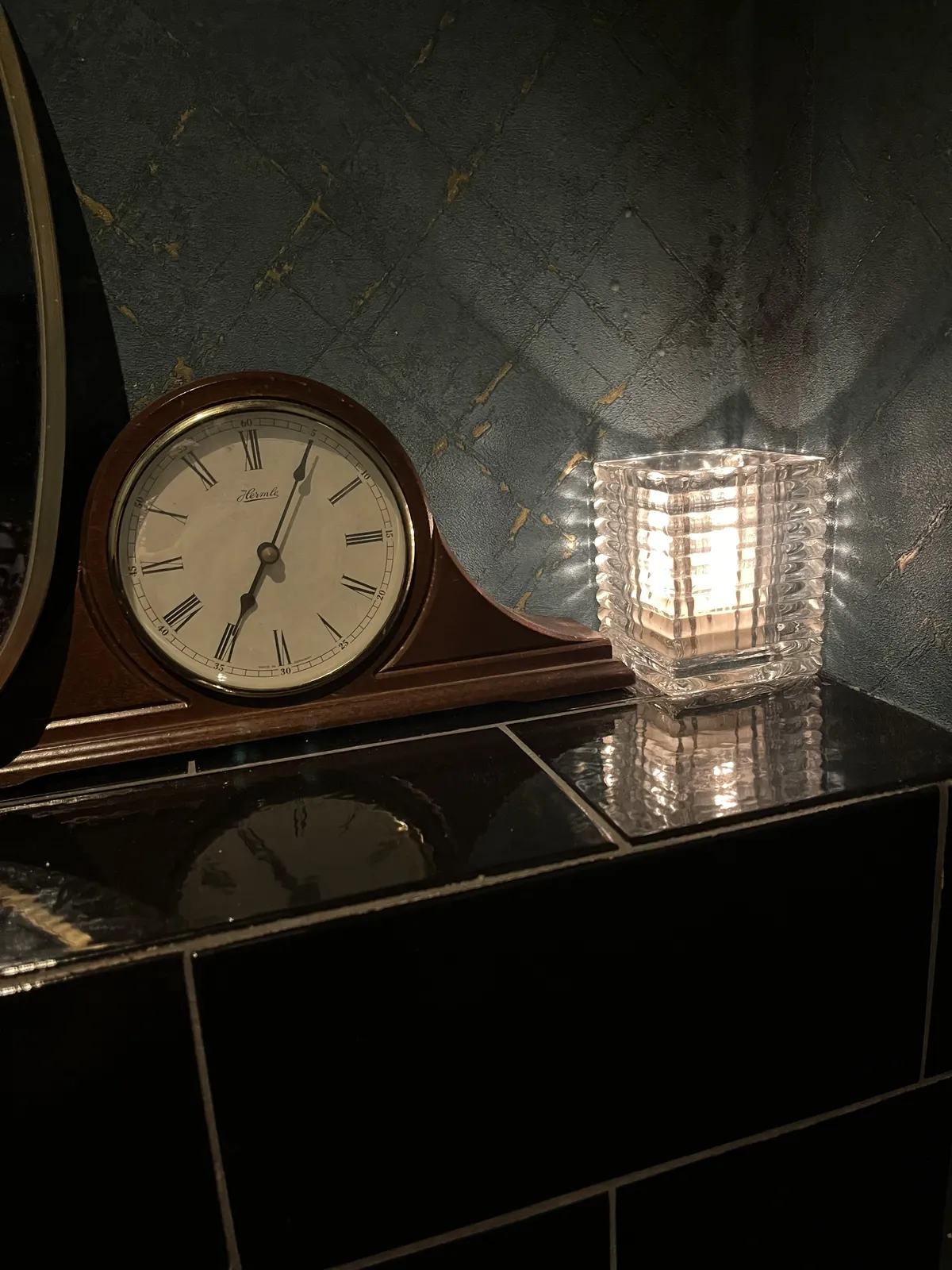 An old, wooden mantel clock on a black tiled shelf, next to a heavy glass lamp. The clock points at 5 past seven.