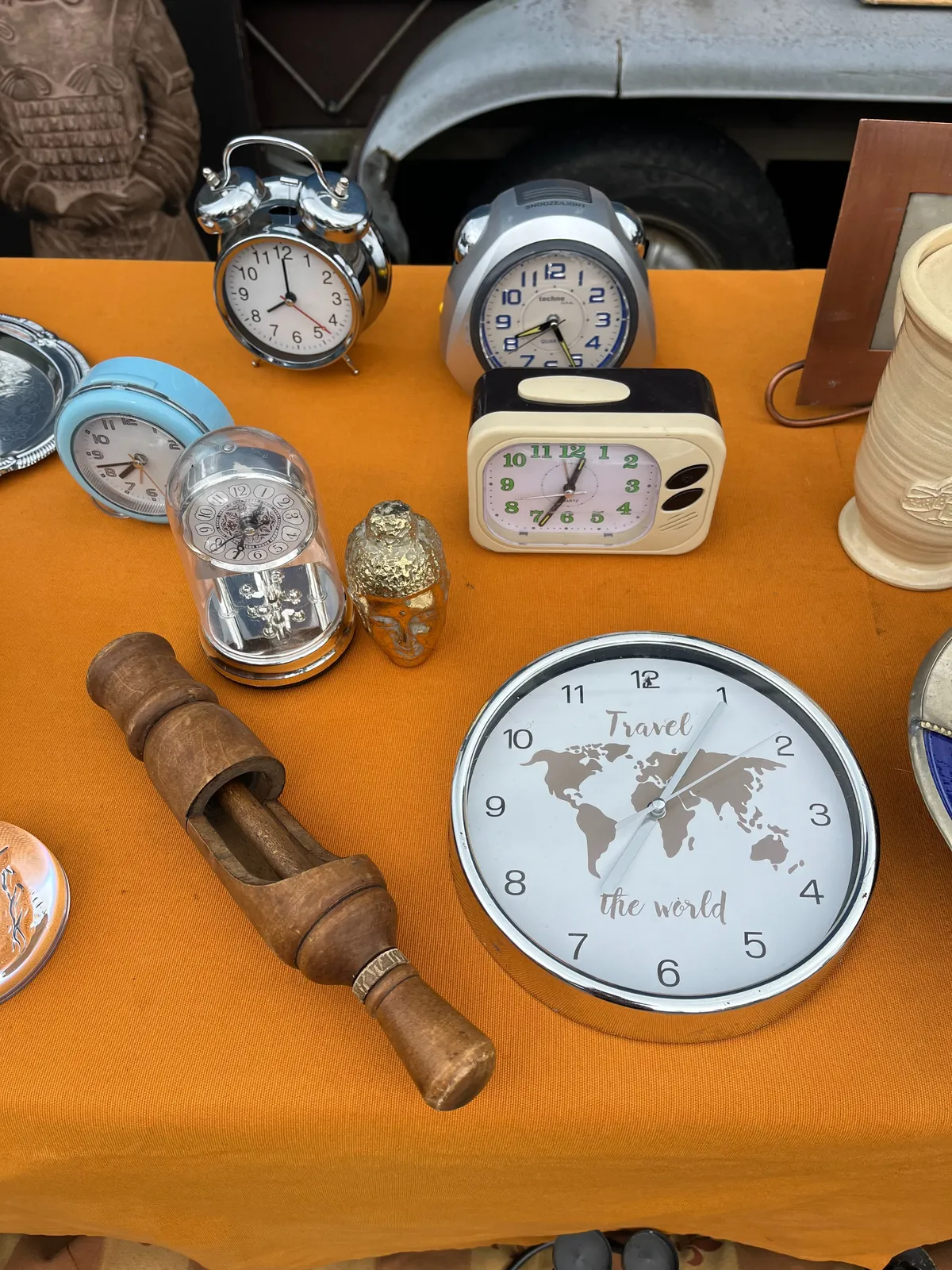 There’s some stuff for sale on this orange rug. Mostly clocks, but also a nut cracker. A black, white and green, rectangular plastic alarm clock with rounded corners points at 34 minutes past 12. The largest clock has a picture of the world with the words, Travel the World, printed on its face. It points at 5 past 7. A little round, blue alarm clock points at 18 minutes to 8. A classic alarm clock, the one with two bells on top, points at 8 o’clock. And finally, a more modern looking alarm clock points at 26 minutes past 8. 