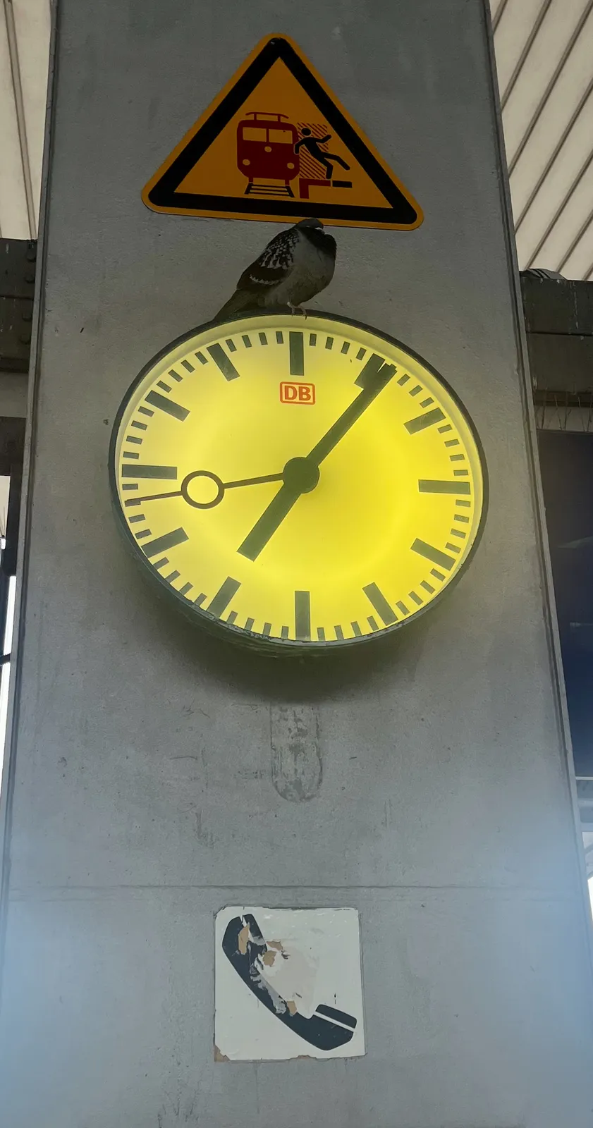 A pigeon sits on top of a clock. Above the bird there’s a warning sign which seems to indicate that you should not fall in front of a train. Below the clock there’s a sticker with an icon of an old fashioned phone horn on it. On the clock there’s a logo of the Deutsche Bahn, the German Railways. The clock points at 6 minutes past seven. 